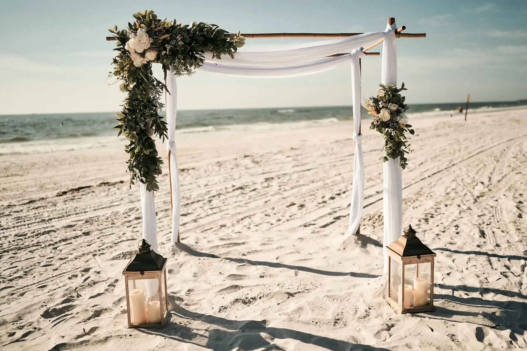 Beach wedding arch with white fabric, flowers, and lanterns.