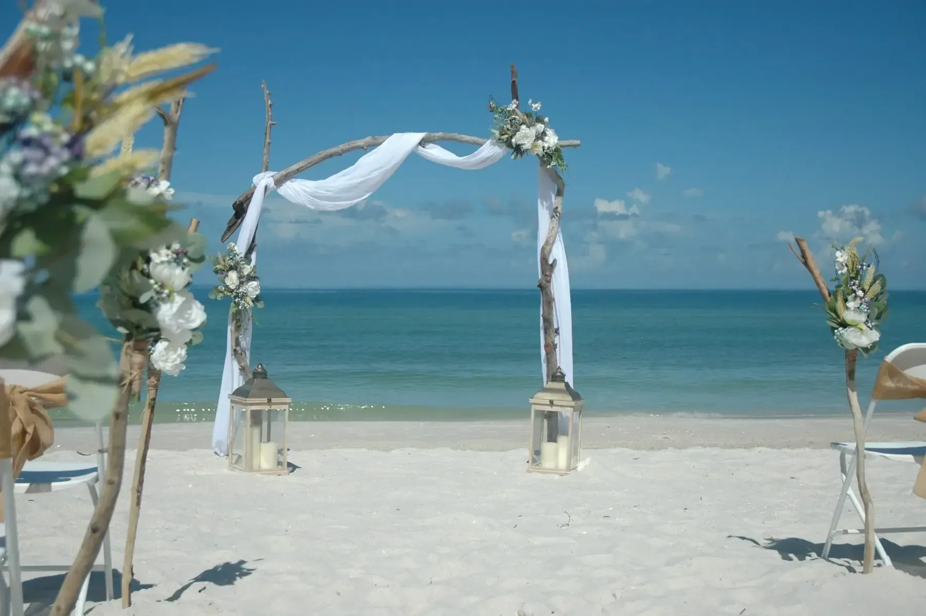 Beach wedding setup, arch decorated with white fabric and flowers, ocean in the background.