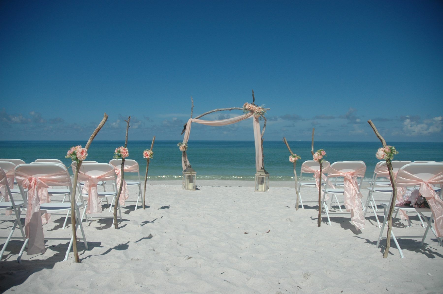 Beach wedding setup with white chairs, driftwood accents, and pink decorations, facing the ocean.