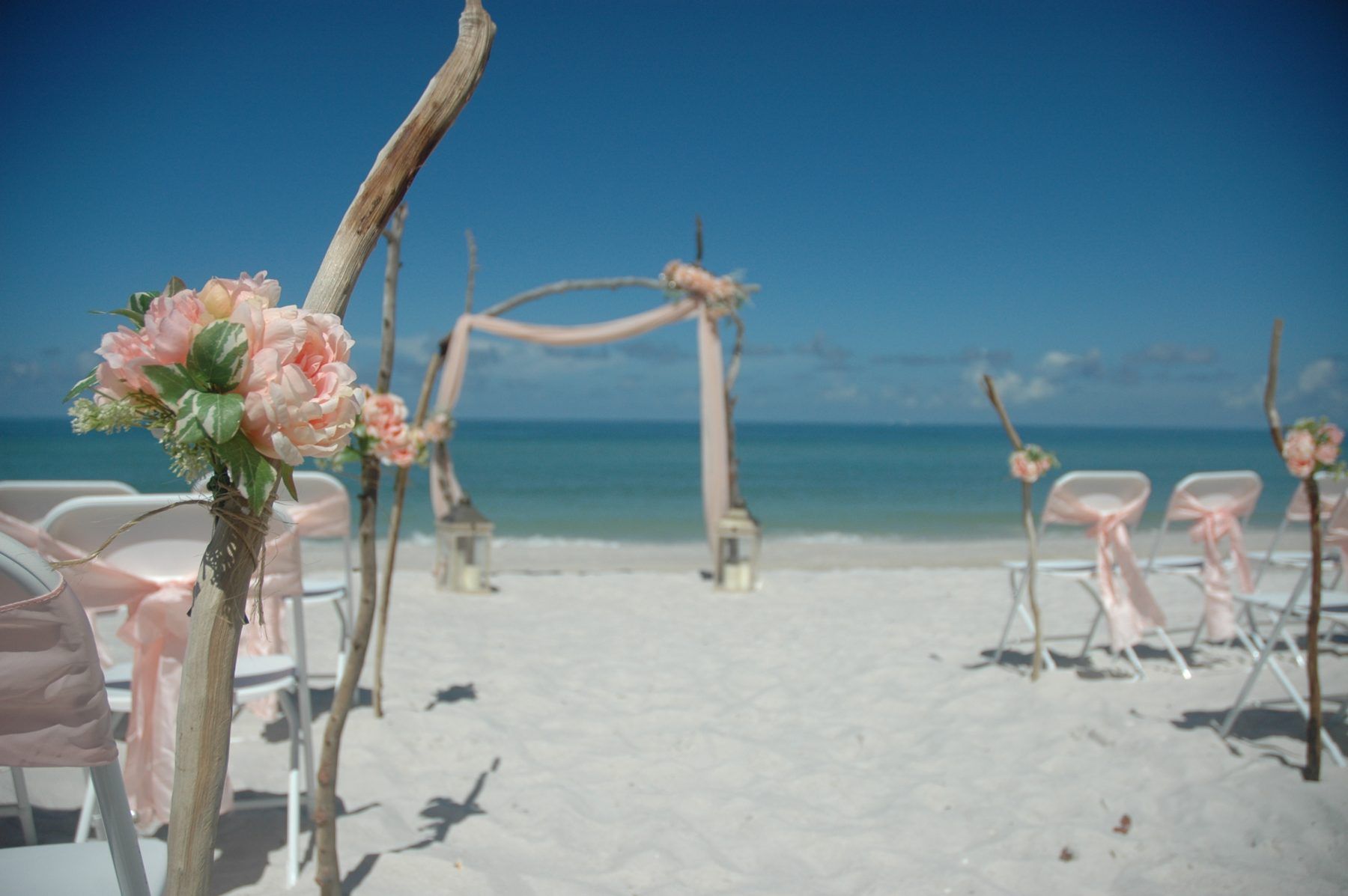 Beach wedding setup: arch, chairs, and decorations on the sand, with ocean and blue sky in background.