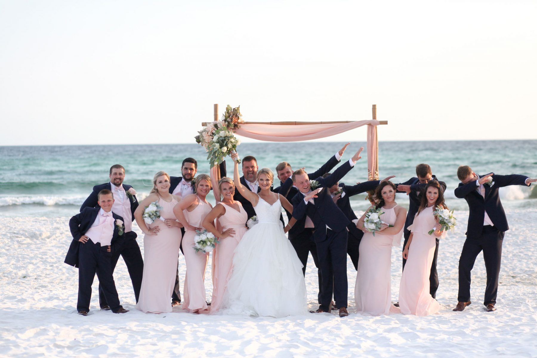 Wedding party on beach, bride in white dress, bridesmaids in pink, groomsmen in black suits, posing by ocean with floral arch.