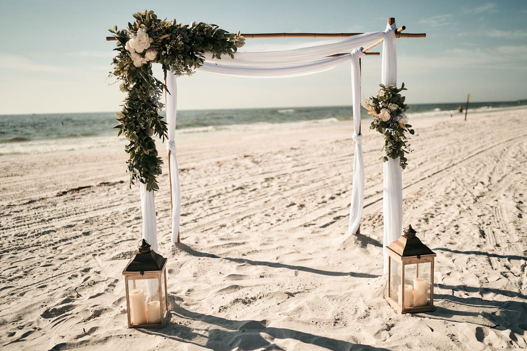 Wedding arch with white fabric and flowers on a beach, flanked by lanterns.