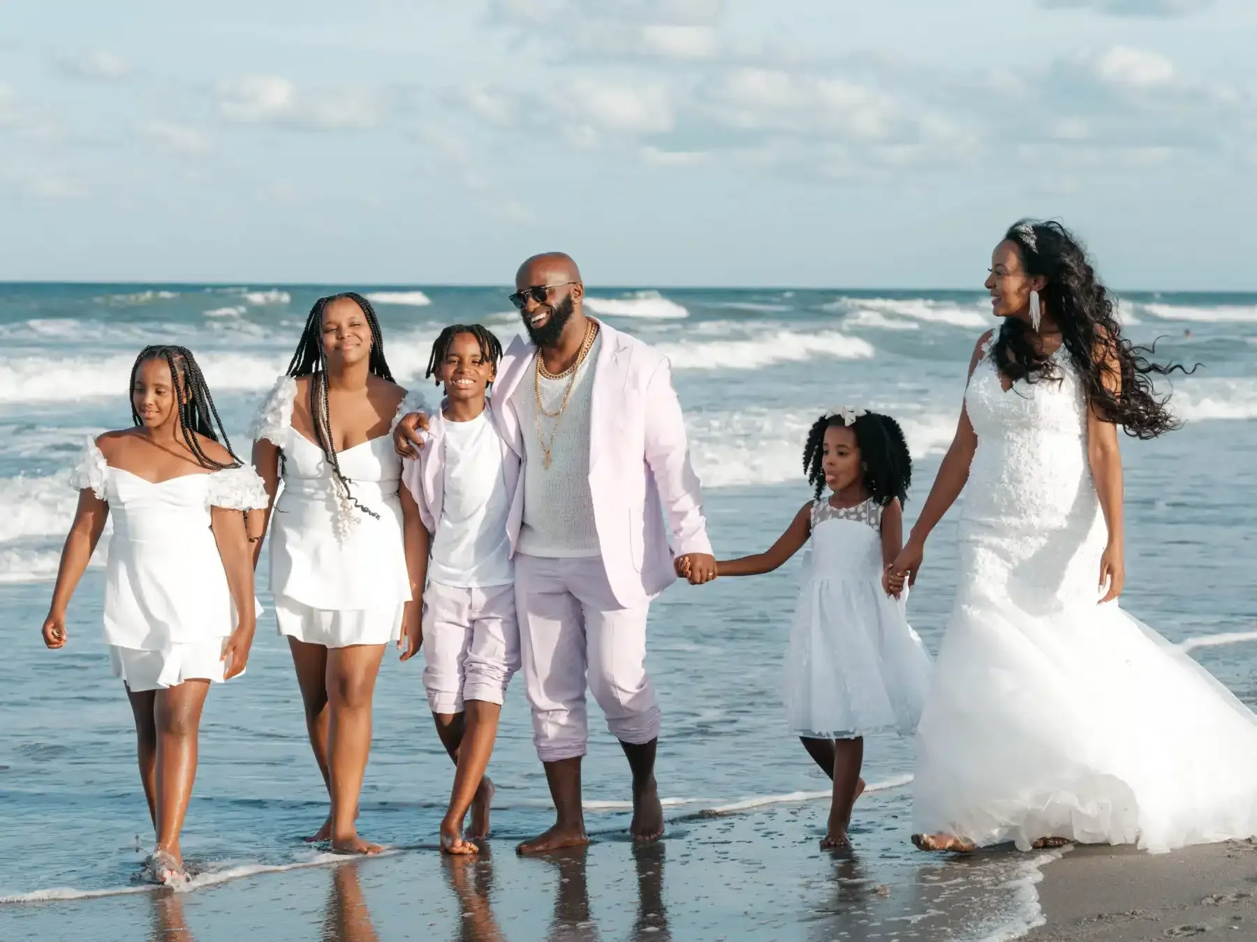 Family of six in white outfits walks on beach; bride in wedding dress.