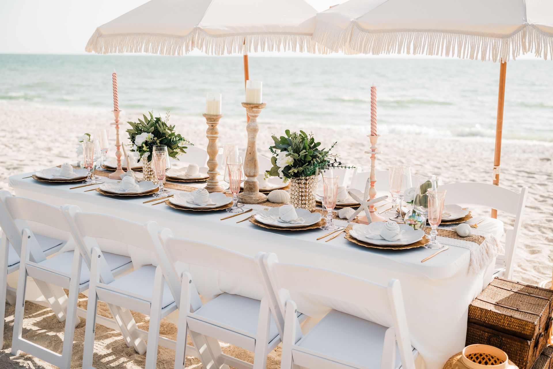 Beachside table setting with white umbrellas, chairs, and elegant decor.