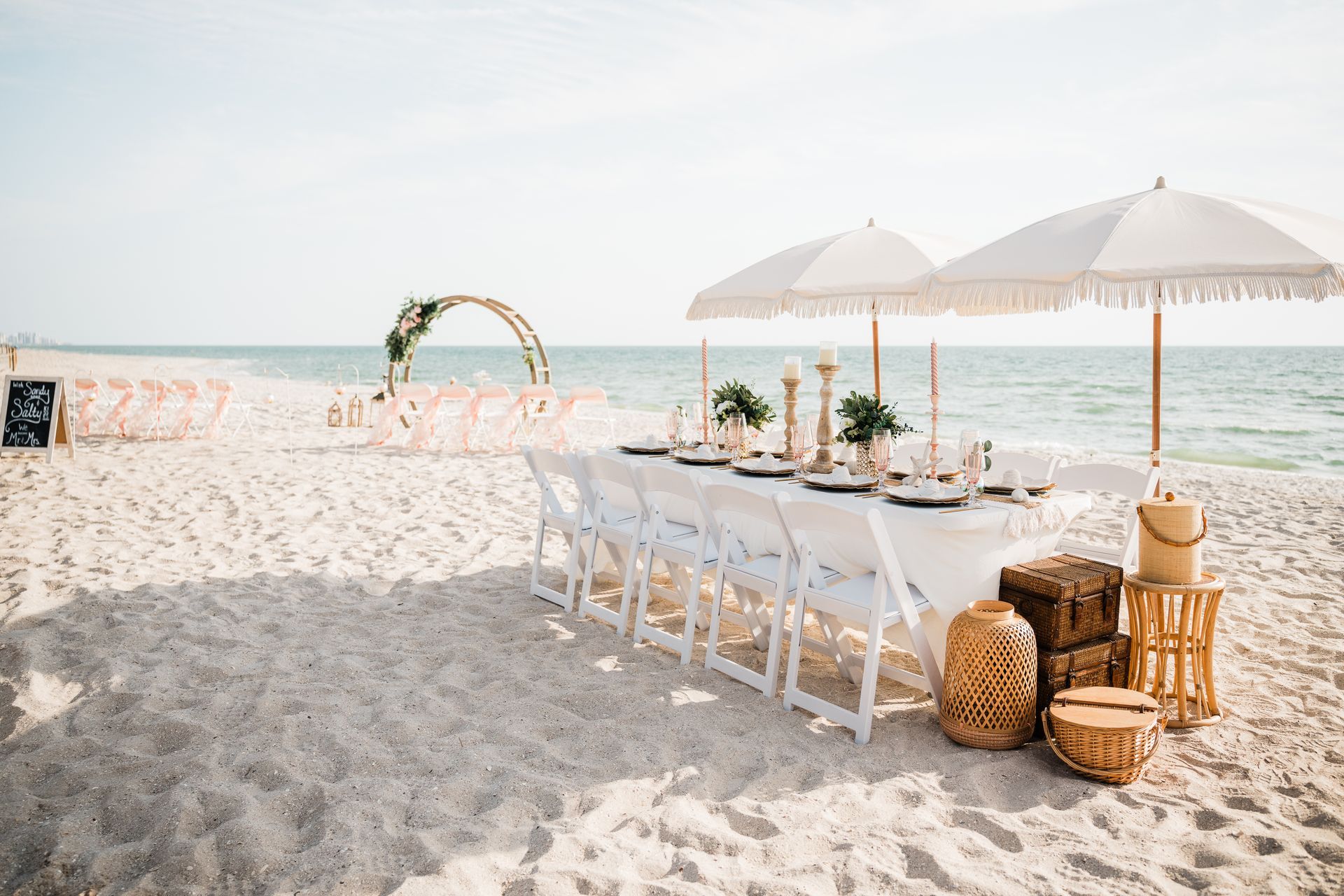 Beach wedding setup with tables, umbrellas, chairs, and arch in the sand.