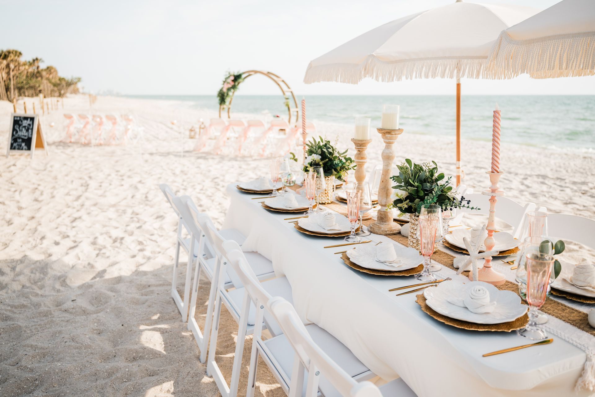 Beach wedding reception setup with white table, gold accents, pink decor, and parasols.