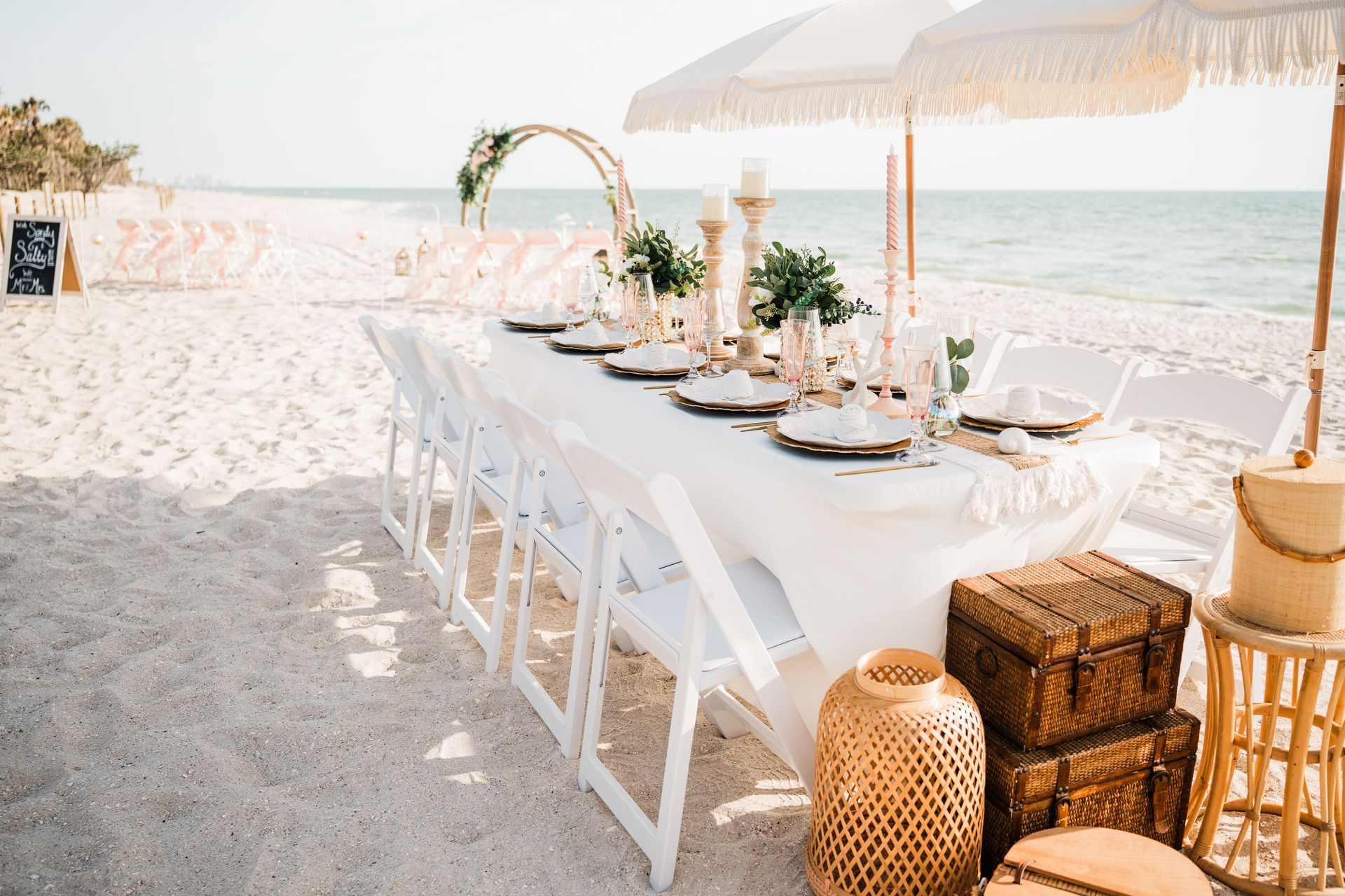 Beach wedding reception setup: long white table, chairs, decorations, ocean backdrop.