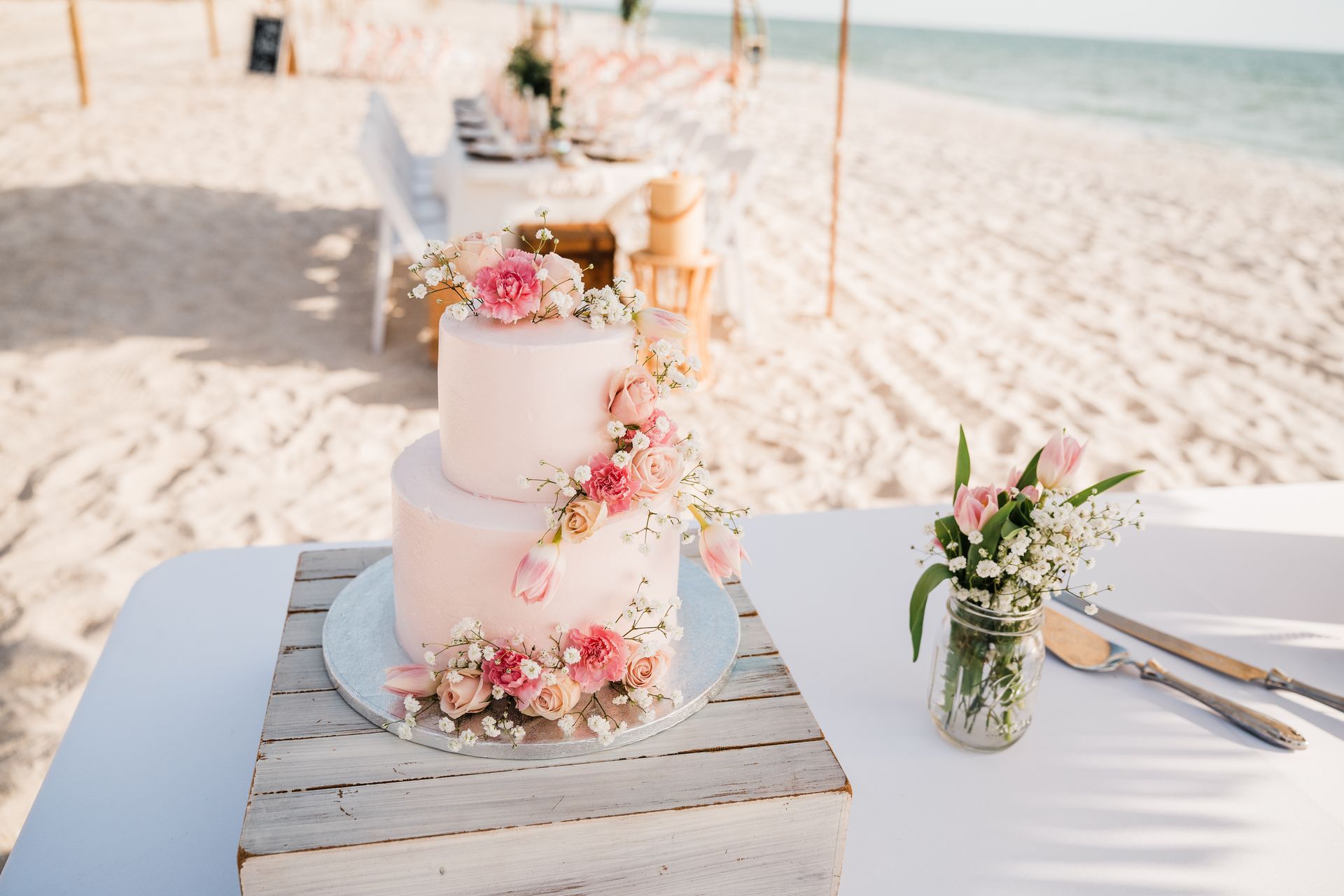 Two-tiered pink wedding cake with flowers on a beach, next to a small flower arrangement and silverware.