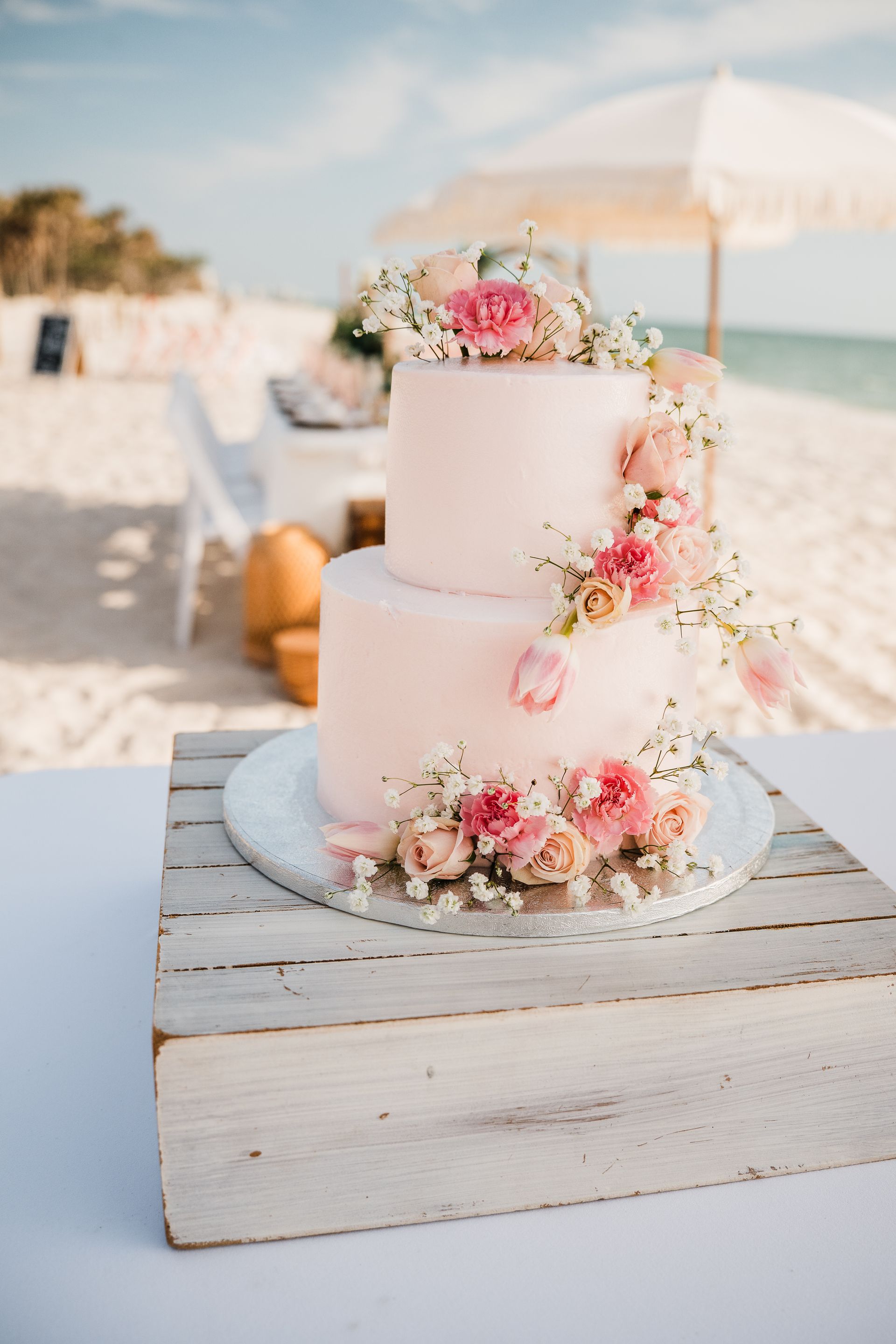 Two-tiered pink frosted cake decorated with flowers, on a rustic wooden stand, beach setting.