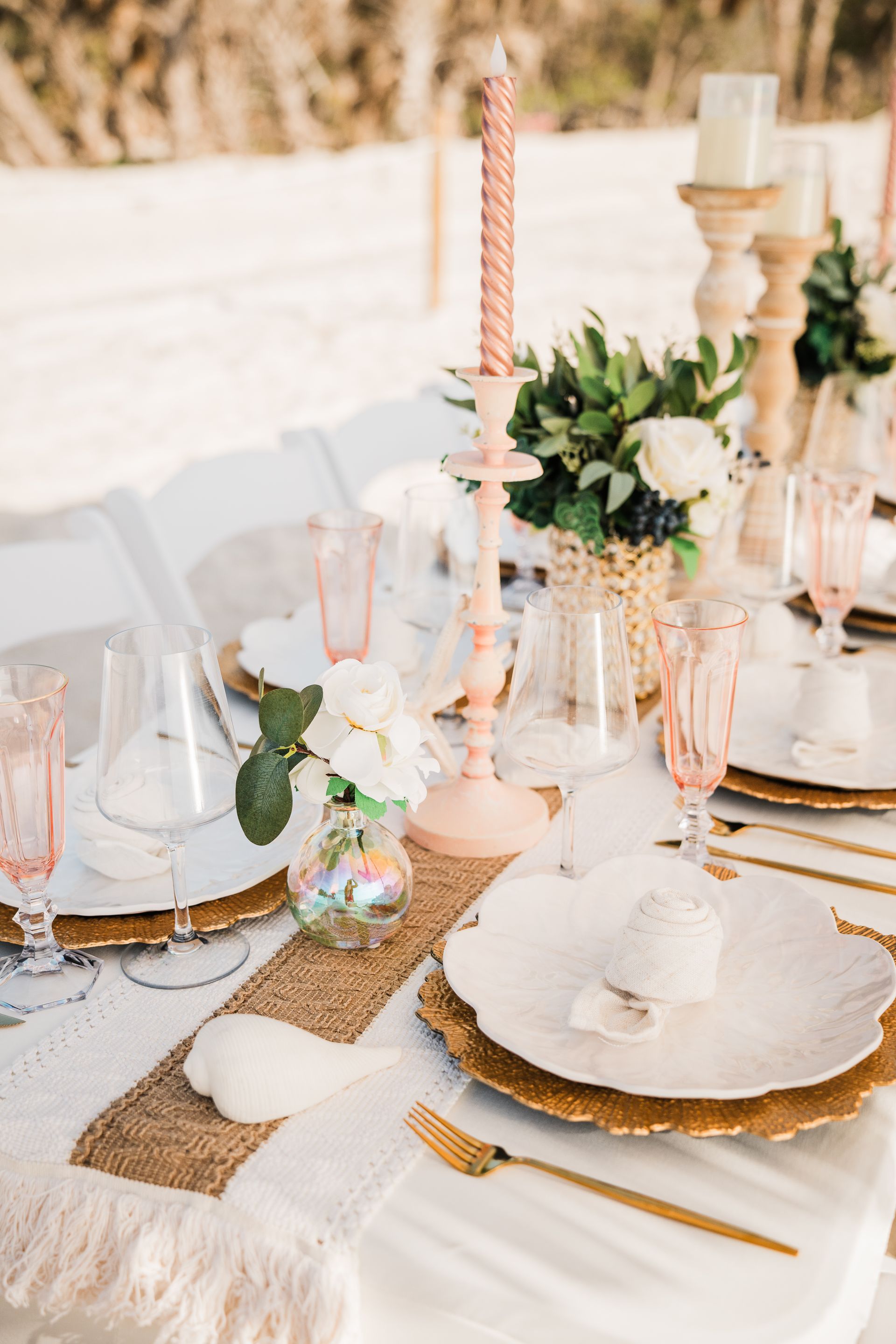 Elegant outdoor table setting with floral arrangements, gold accents, and pink glasses.