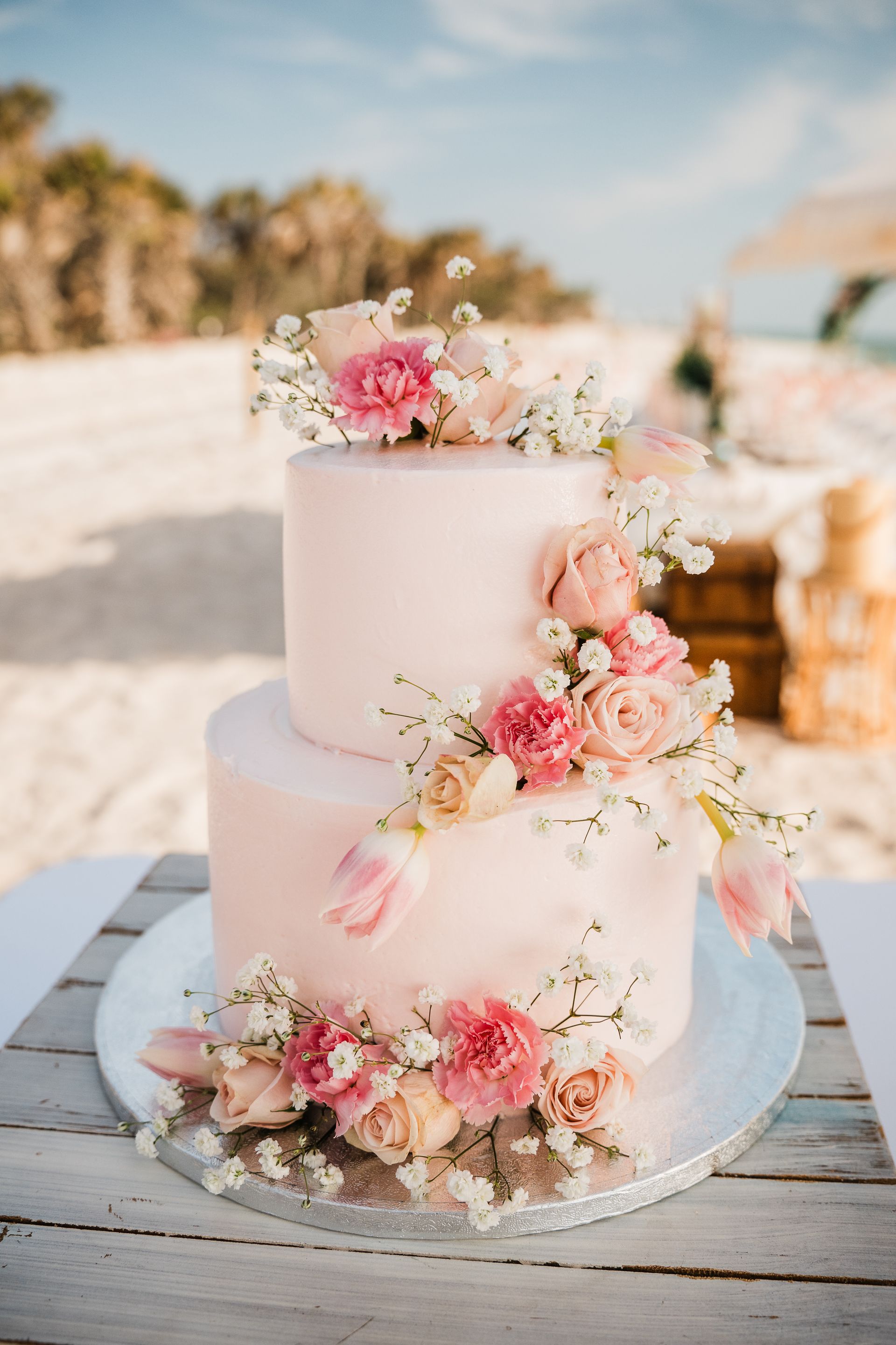 Two-tiered pink frosted cake decorated with flowers, on a beach.