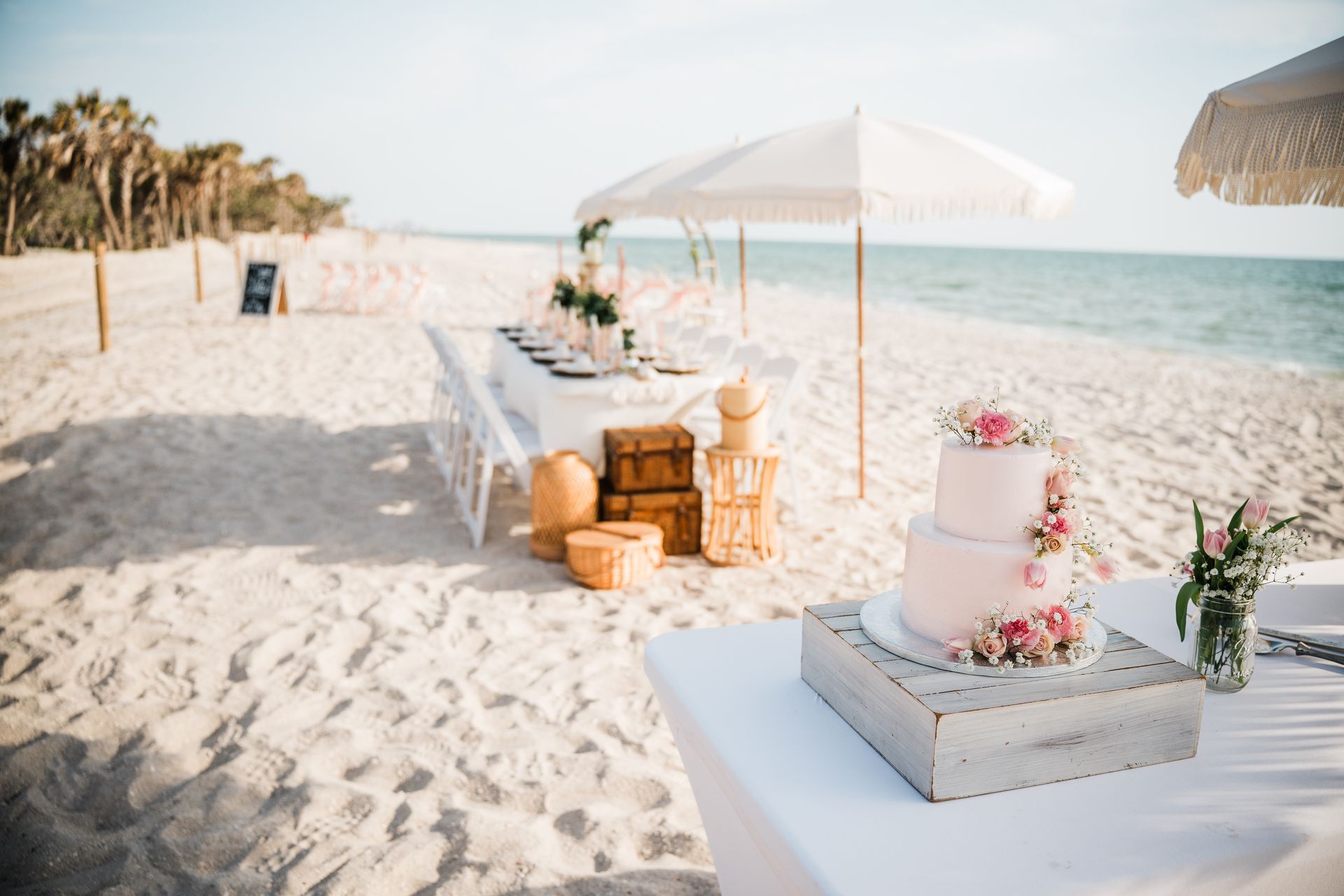 Beach wedding reception setup with cake, long table, and umbrellas.