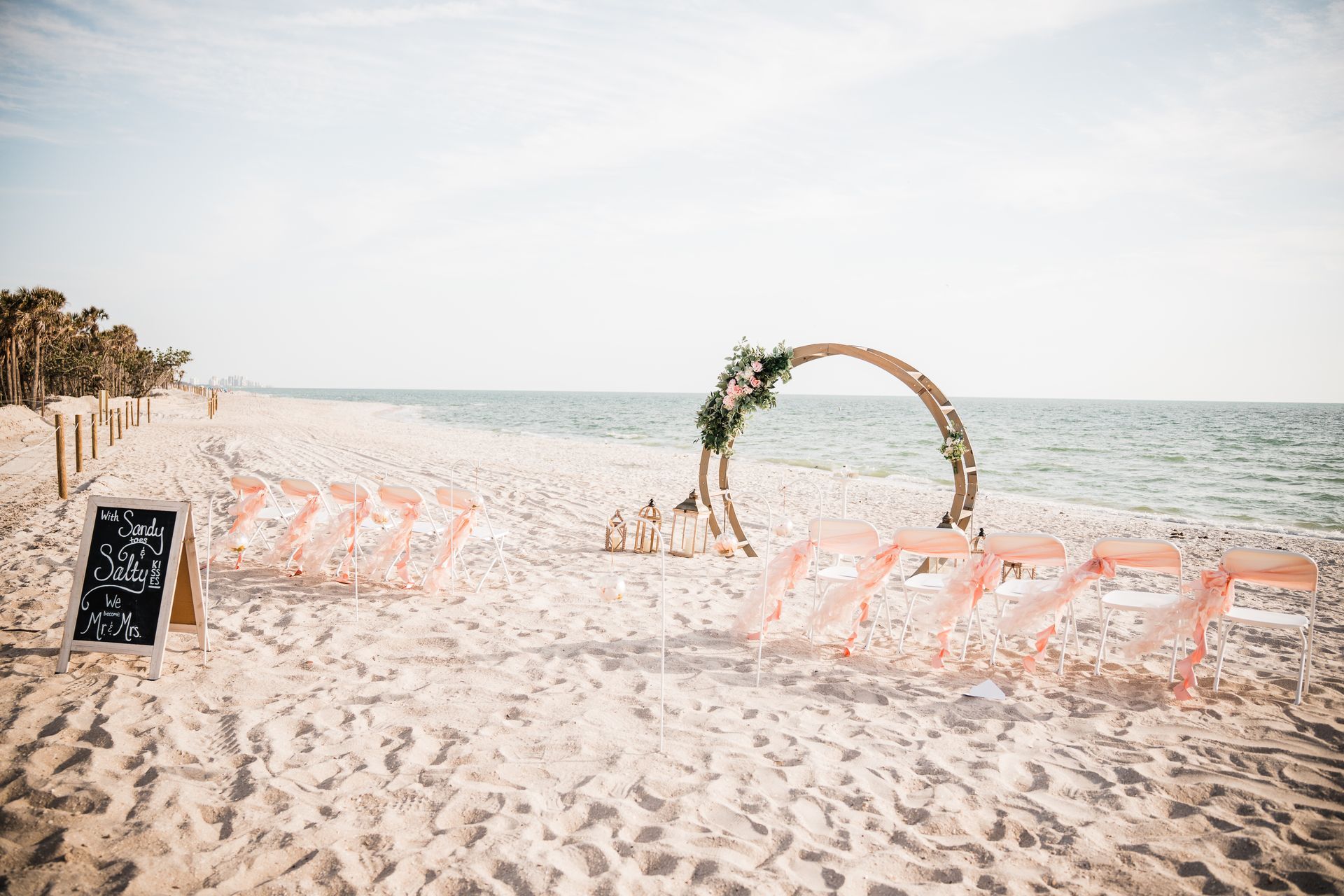 Beach wedding setup with chairs facing a floral arch over the water.