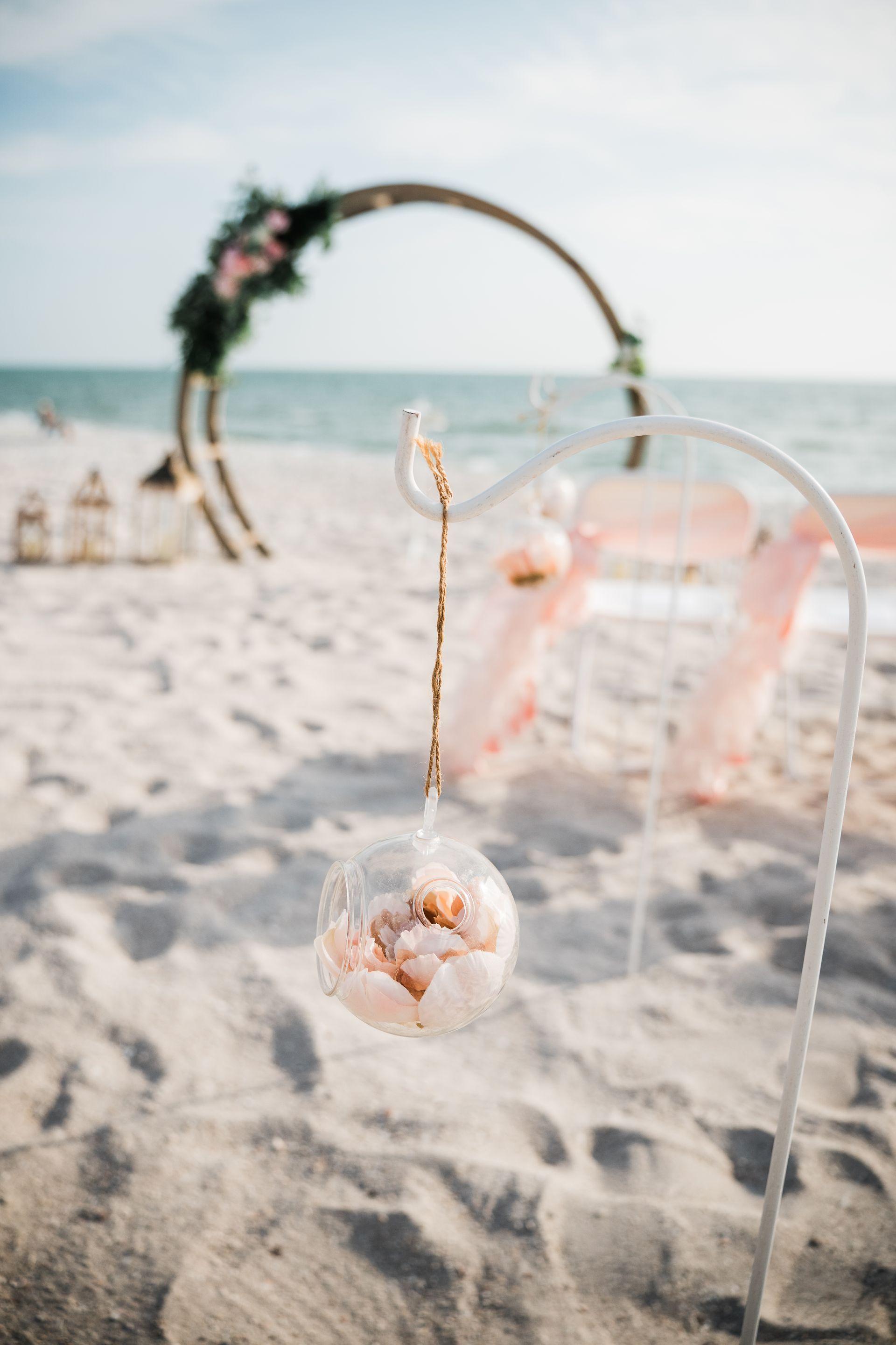 Beach wedding decor: glass orb with flowers hangs from white stand, arch in background.