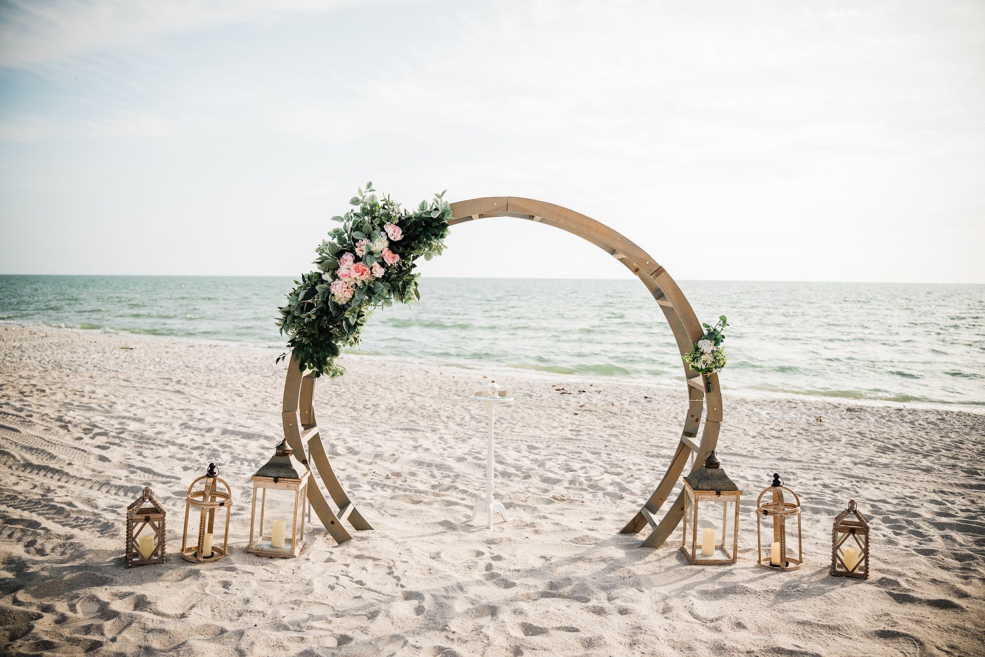Wedding arch on a sandy beach decorated with flowers and lanterns; ocean in background.