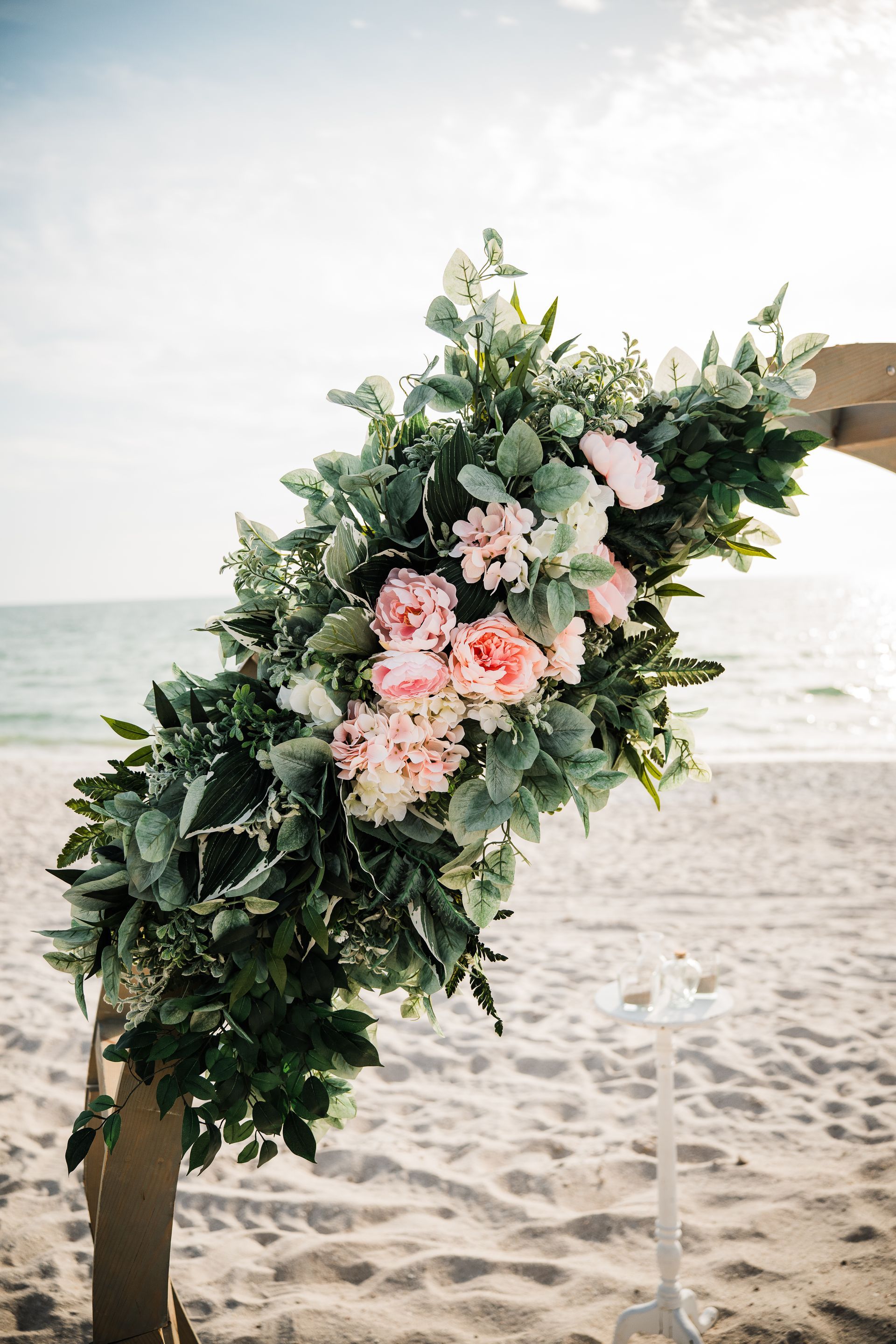 Floral arch decorated with pink flowers and greenery on a beach with the ocean in the background.