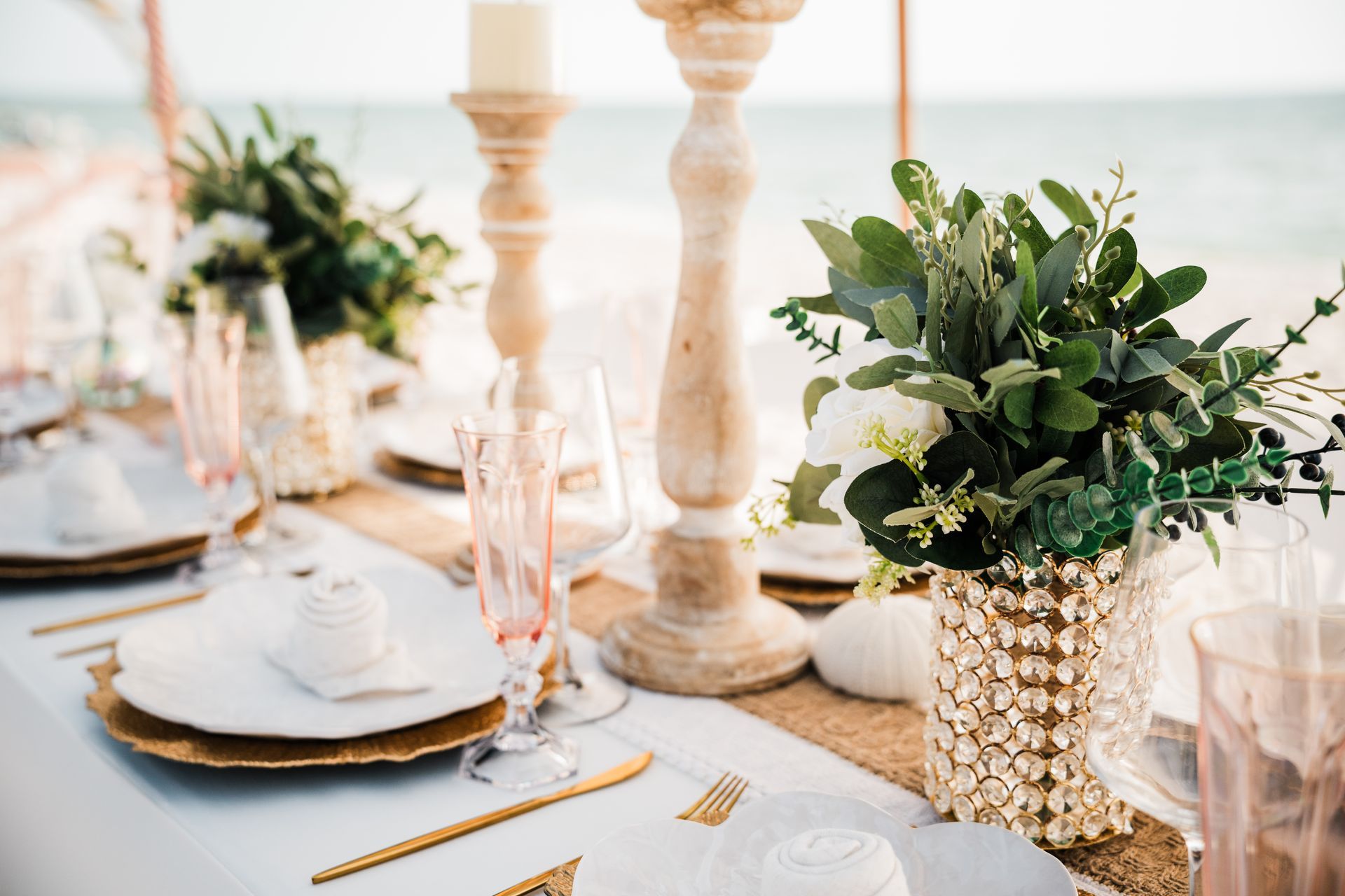 Beachfront table setting, elegant decor with floral arrangements, candle holders, and gold accents.