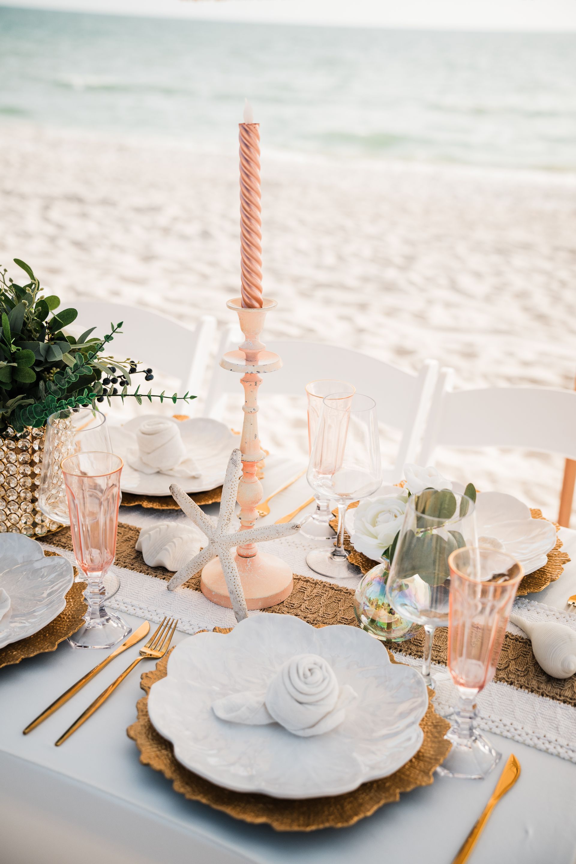 Beach table setting with gold accents, pink candle, and white dishes.