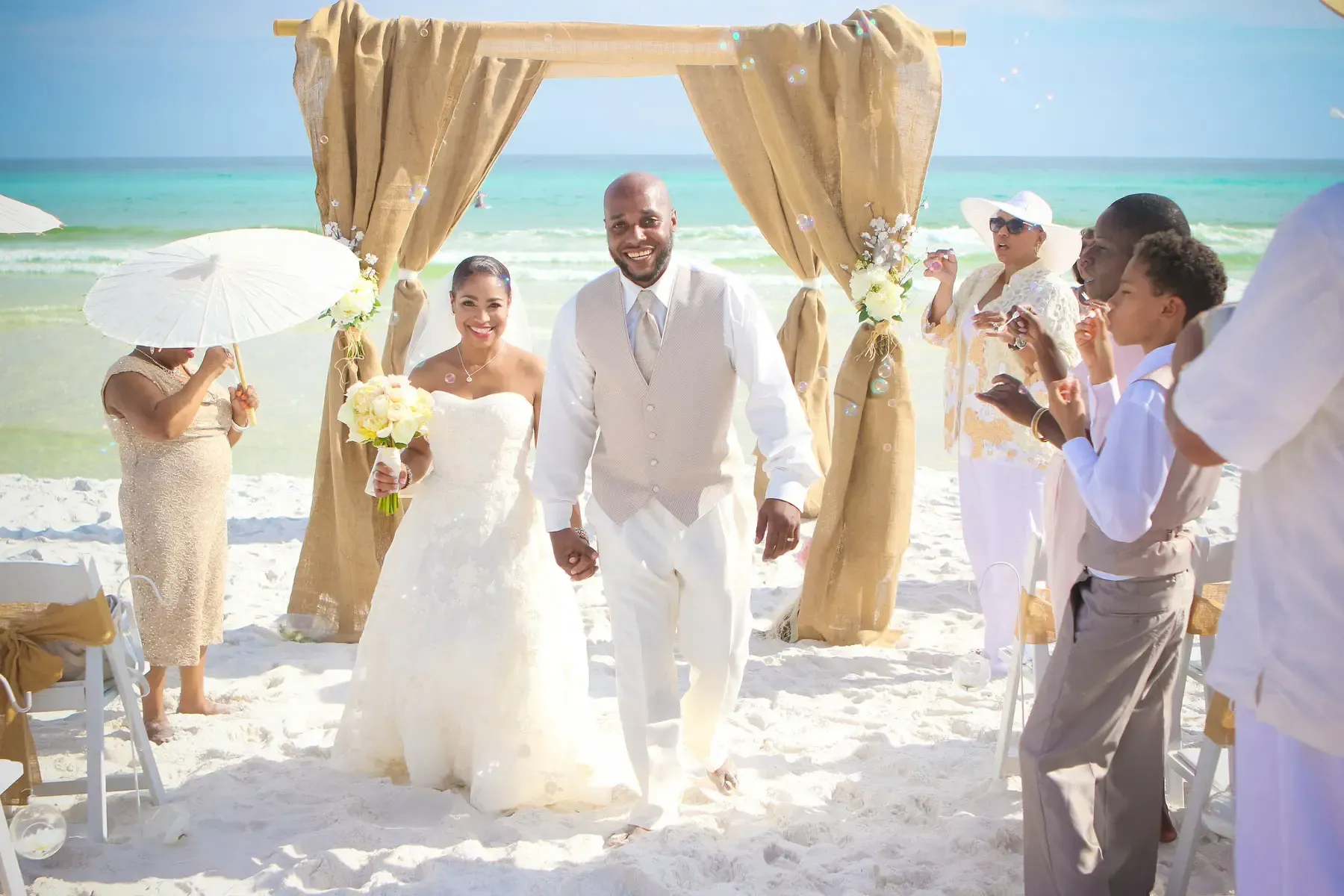 Newlyweds exit beach wedding ceremony under decorated archway.