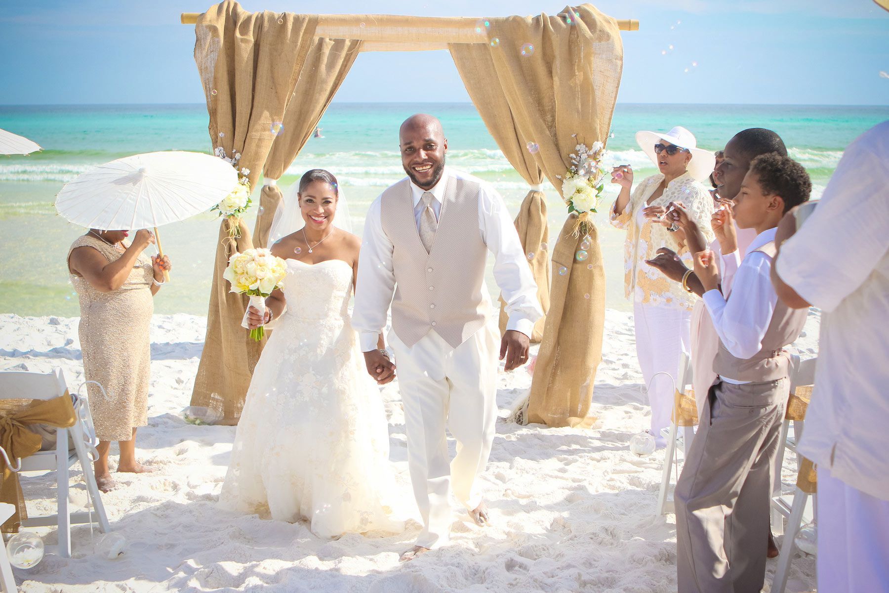 Couple walking down the aisle after a beach wedding; white sand, turquoise water, wedding arch.