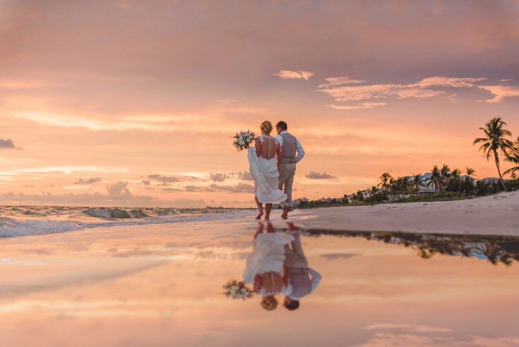 A bride and groom are walking on the beach at sunset.