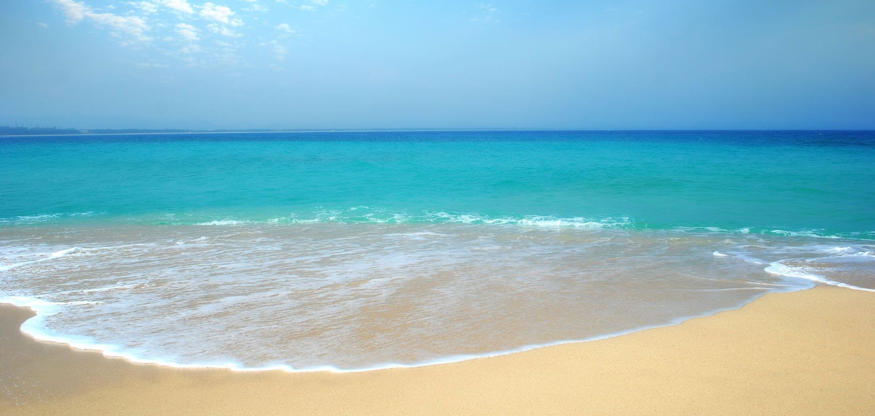 View of a turquoise ocean meeting a tan sandy beach under a blue sky.