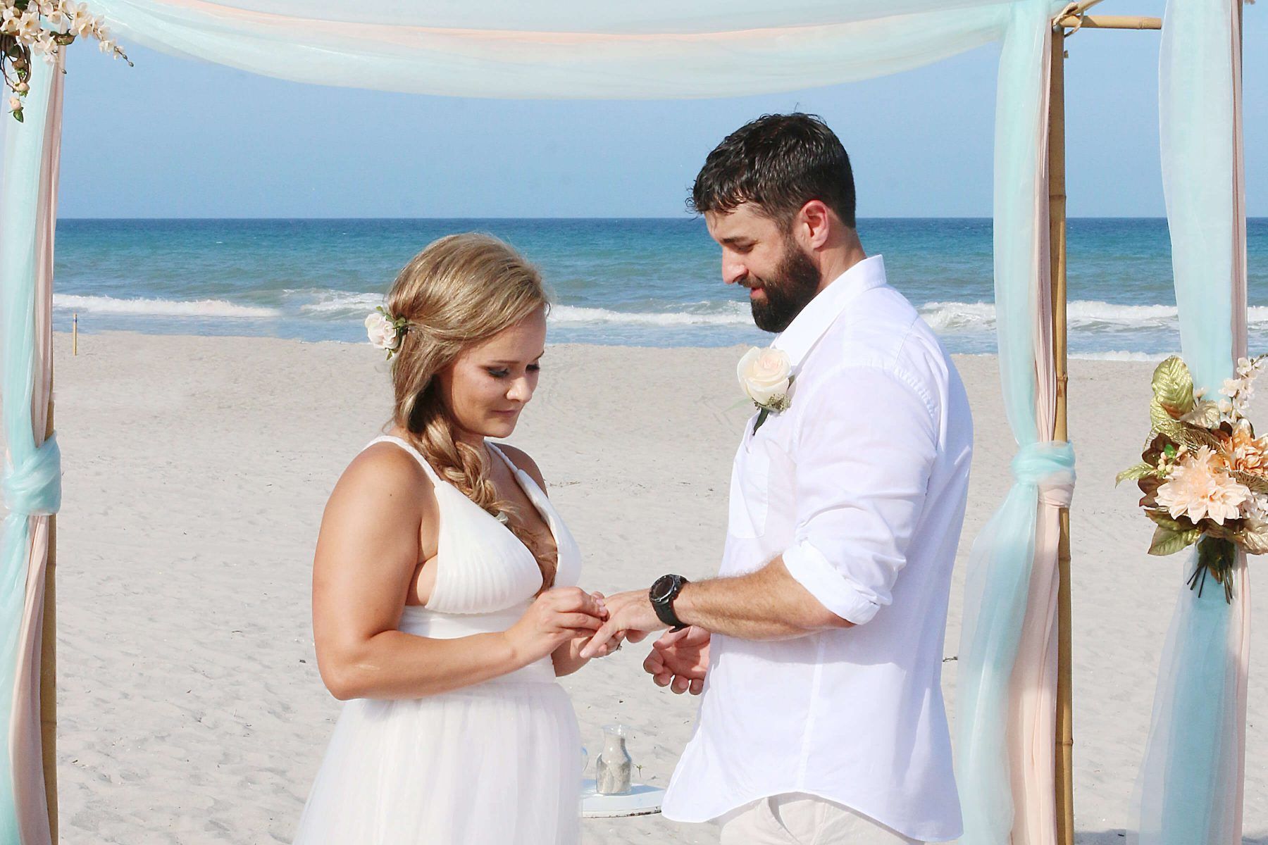 Bride placing a ring on groom's finger during beach wedding ceremony; ocean in background.