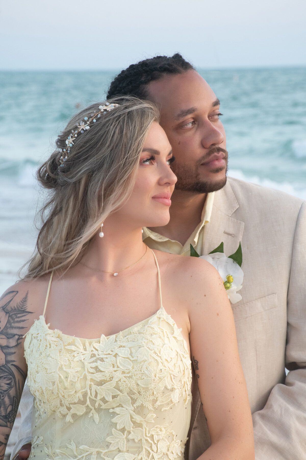 Couple on a beach, woman in lace dress, man in blazer, looking toward the ocean.