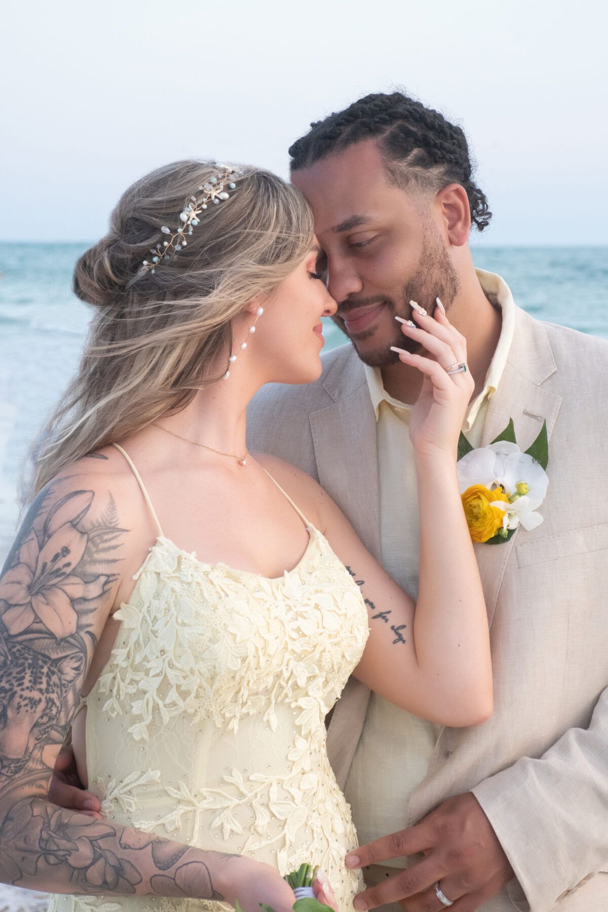 Couple embraces on beach at sunset; bride in lace dress, groom in tan suit.