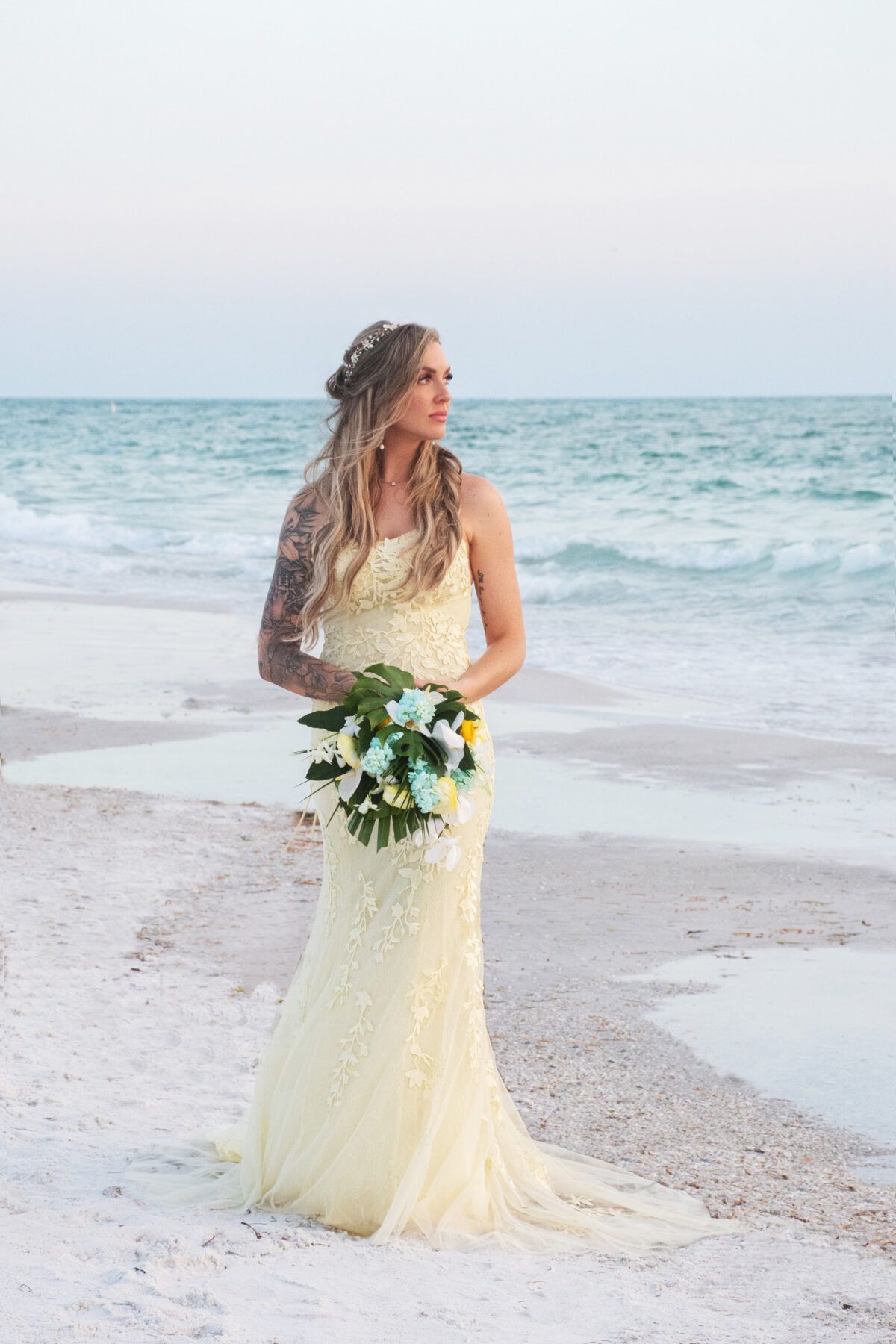 Bride in a pale yellow beaded gown holding a bouquet, standing on a beach, looking out at the ocean.