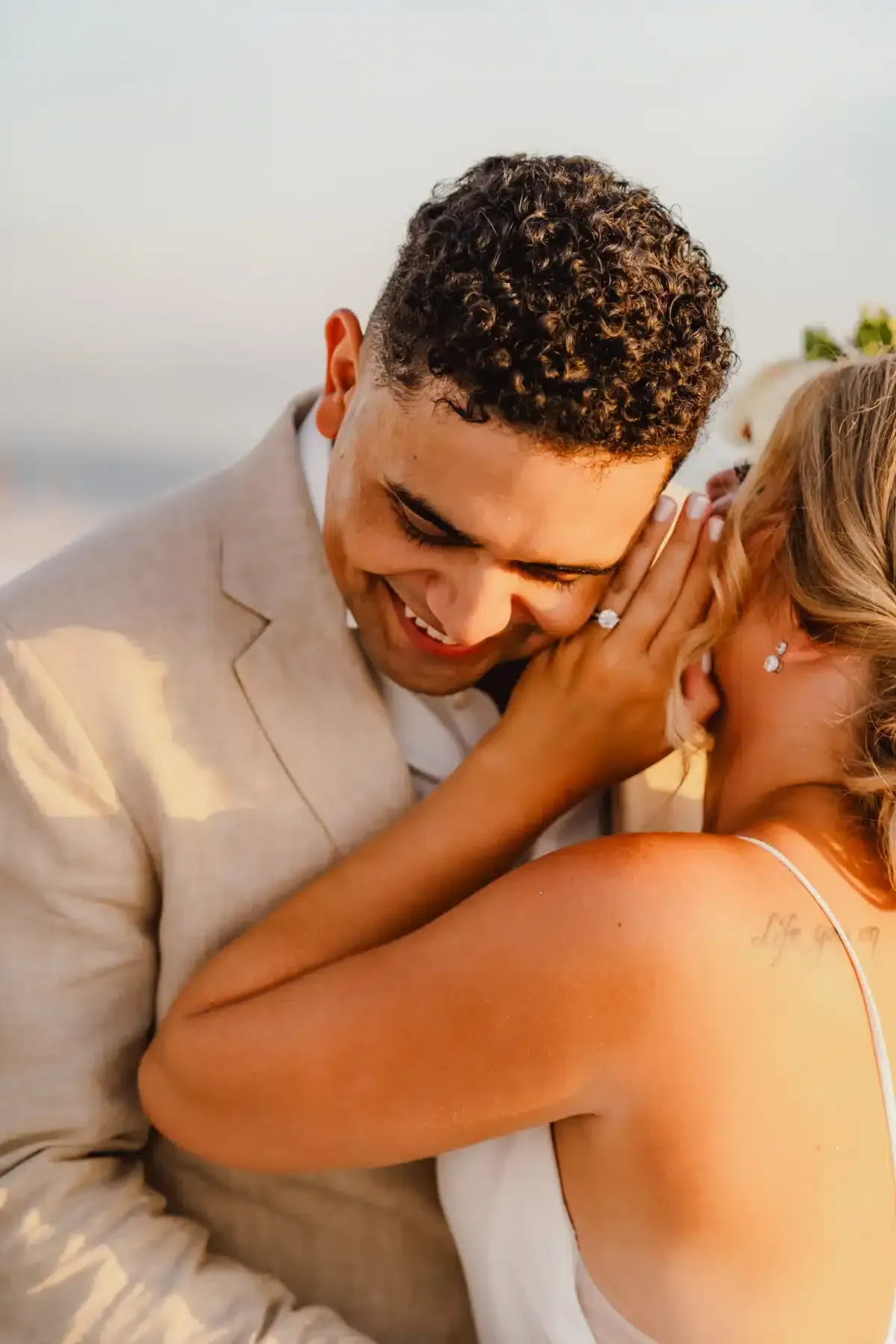 Man in beige suit smiles as woman whispers in his ear. Beach setting, soft light.