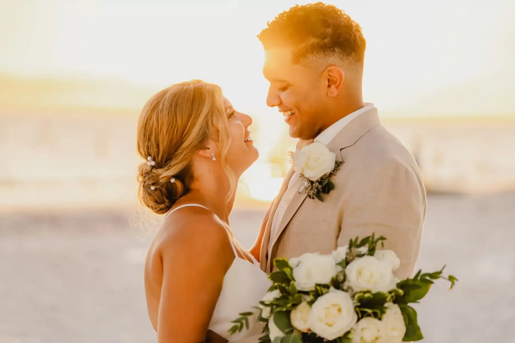 Bride and groom smiling at each other at sunset on a beach; she wears a white dress, he a tan suit.