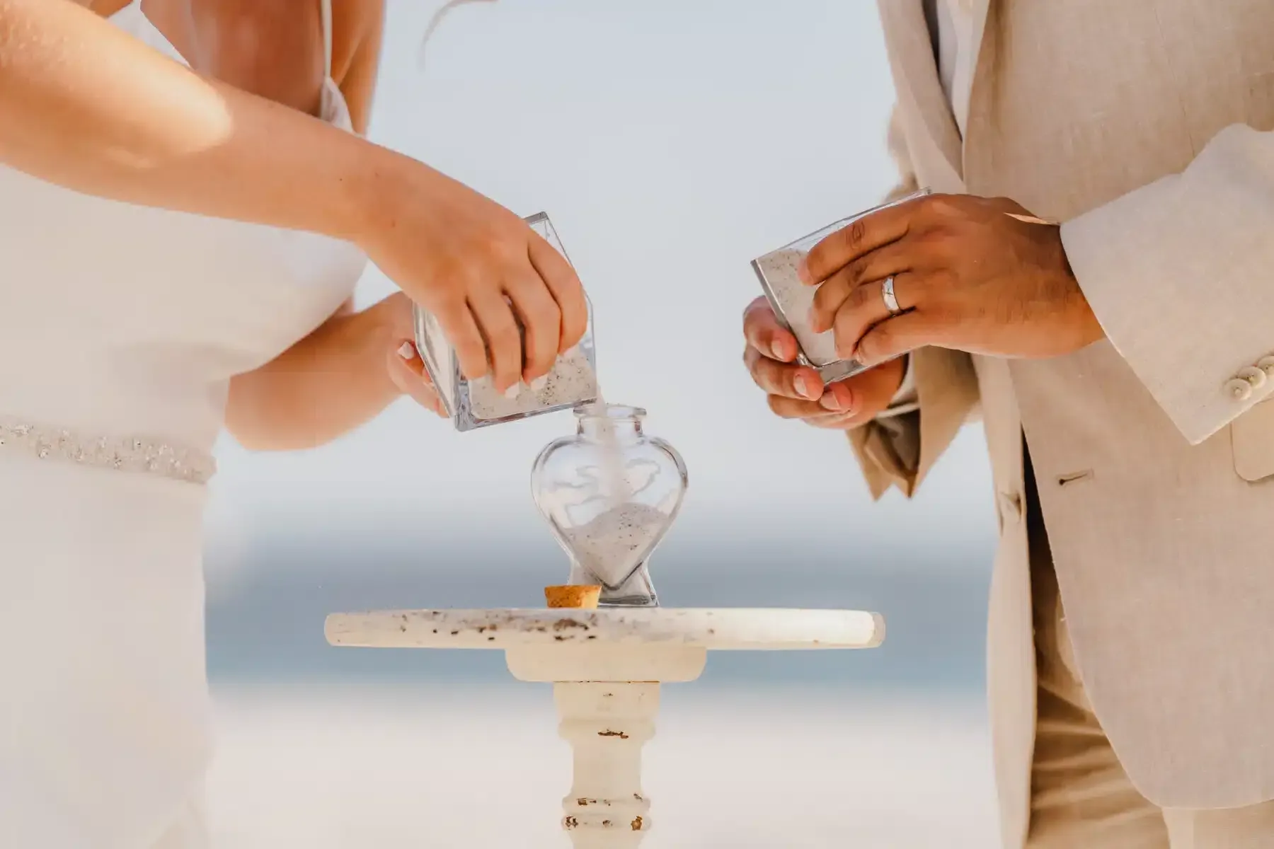 Couple pouring sand into a heart-shaped glass during a beach wedding ceremony.