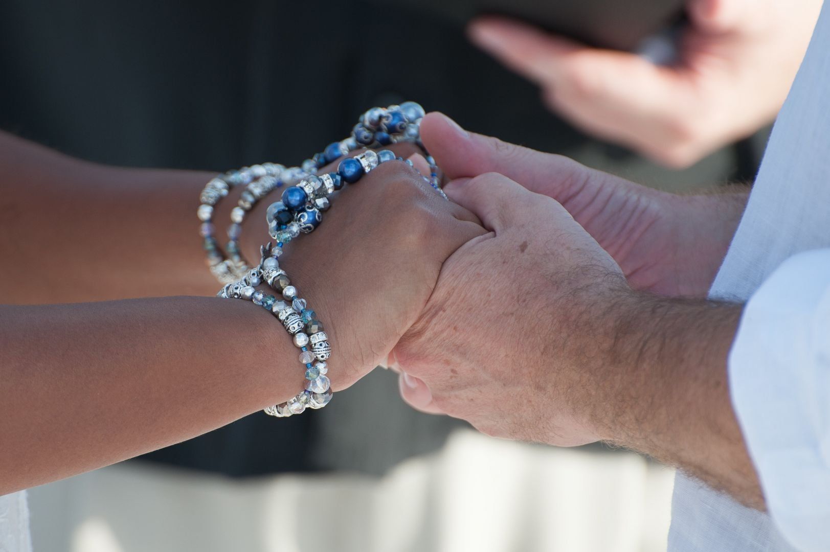 Couple's hands clasped during a ceremony; one wrist adorned with silver and blue bracelets.