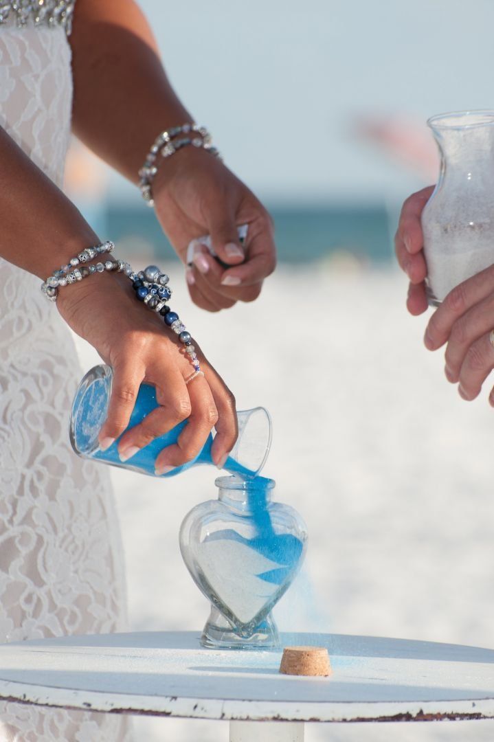Wedding sand ceremony: Bride pours blue sand into a heart-shaped vase with white sand, beach background.