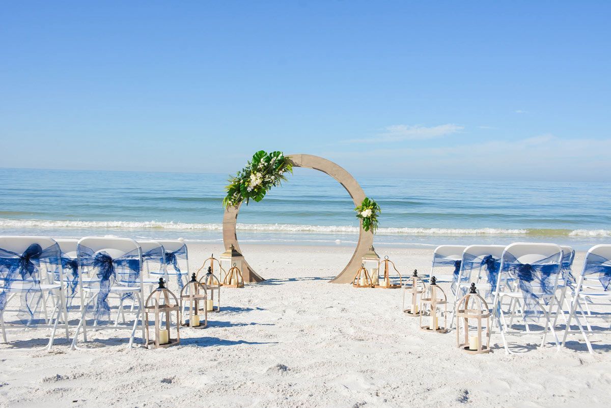 Wedding setup on a beach: circular arch decorated with flowers, chairs with blue sashes, lanterns, ocean in the background.