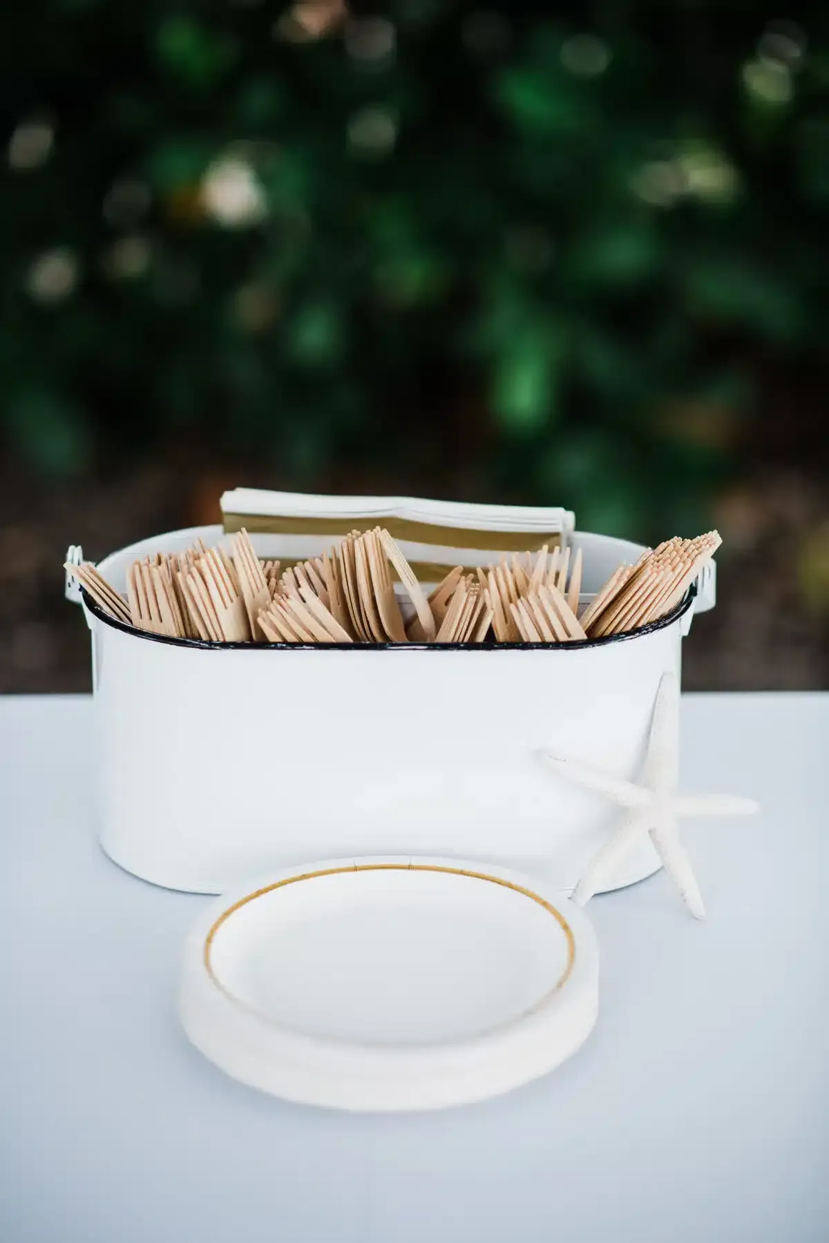 White enamelware container holding wooden cutlery and napkins, with a plate and starfish.