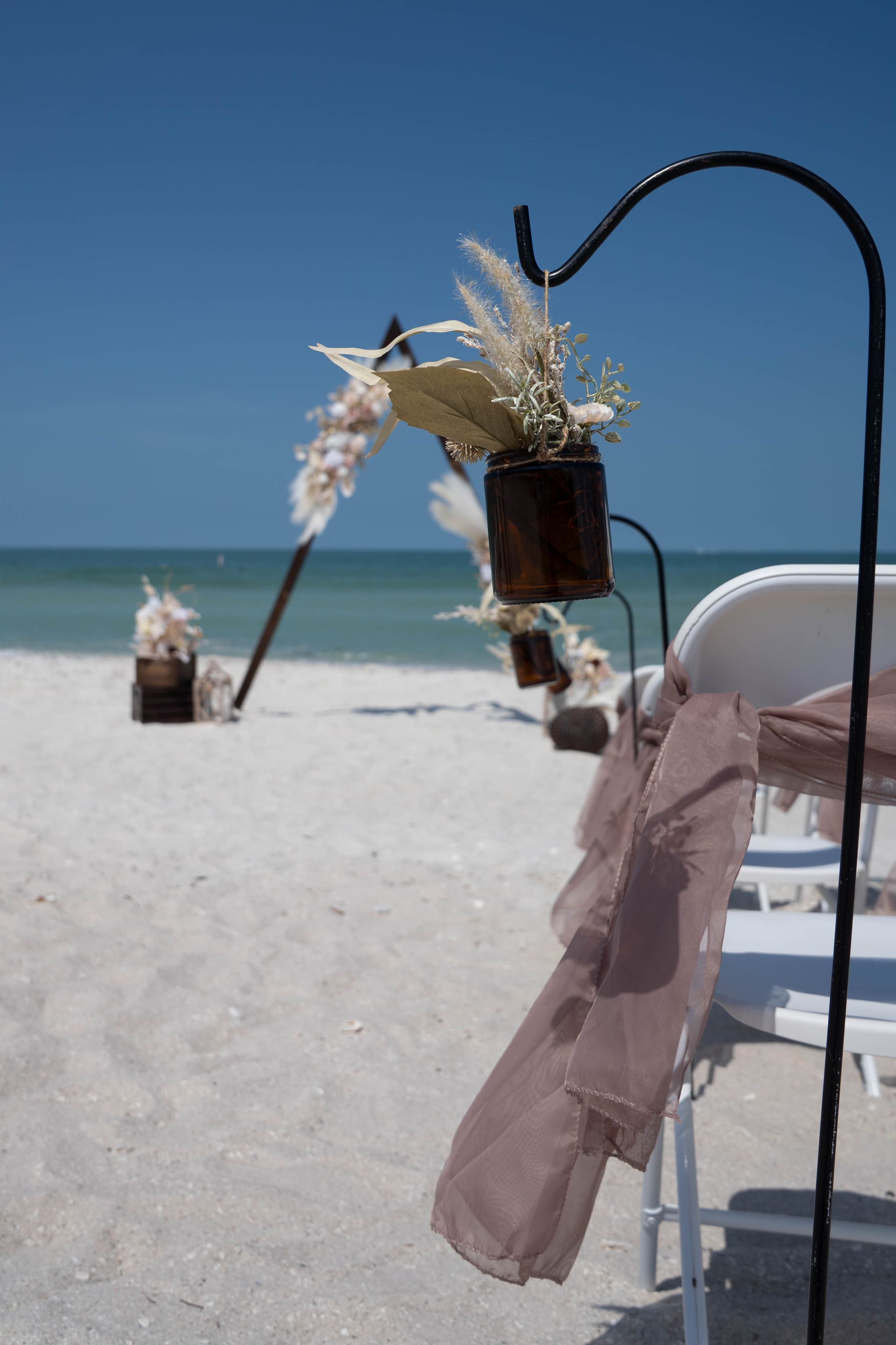 Beach wedding setup with chairs, brown fabric, and floral decorations. Blue sky and ocean in background.