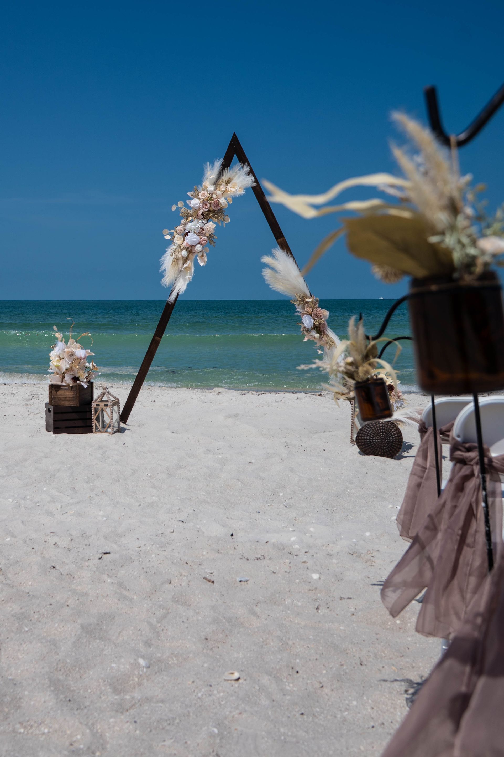 Beach wedding setup: triangular arch with floral decorations, wooden crates, and chairs; ocean background.