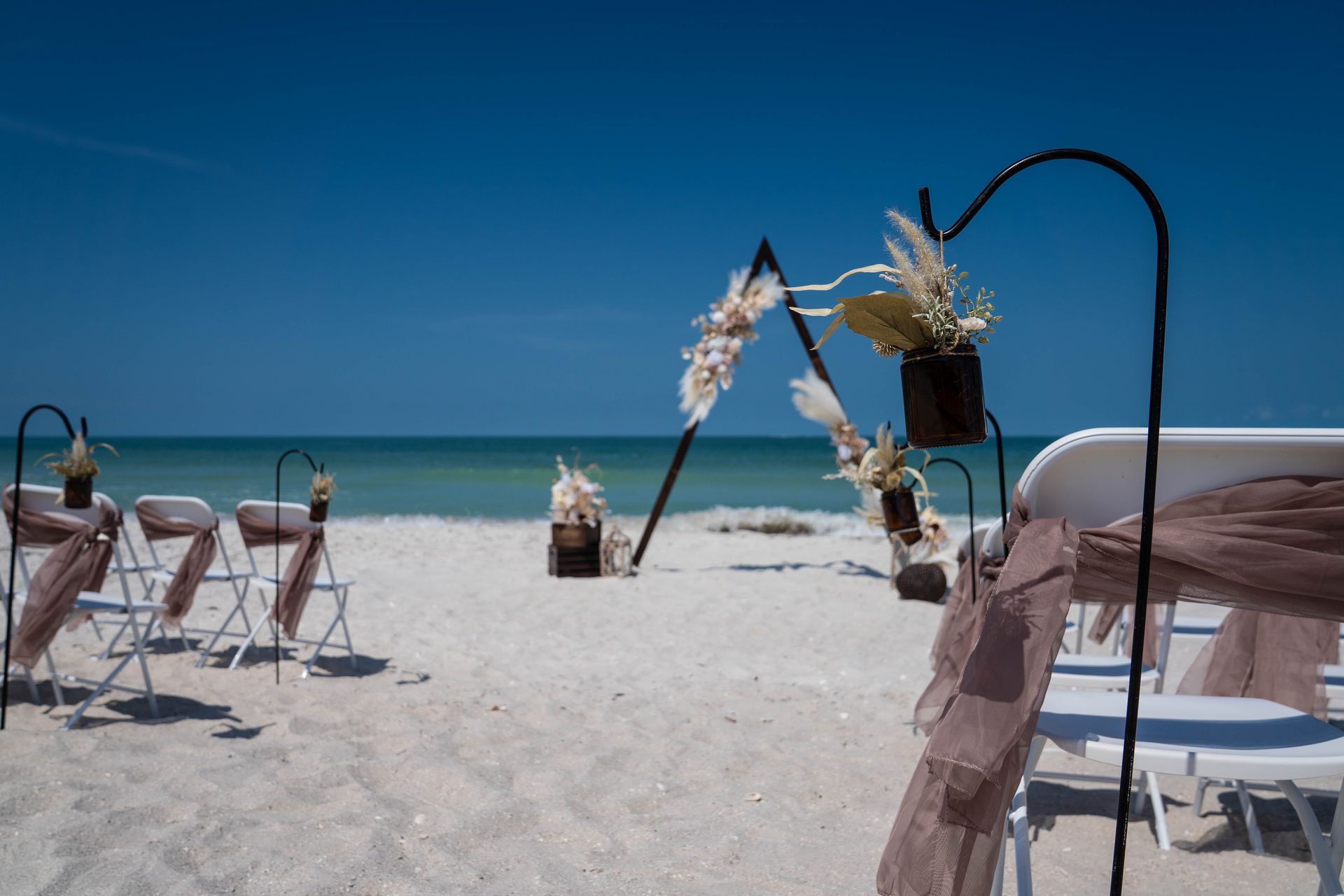Beach wedding setup: chairs with draping, floral arch, ocean, and clear blue sky.