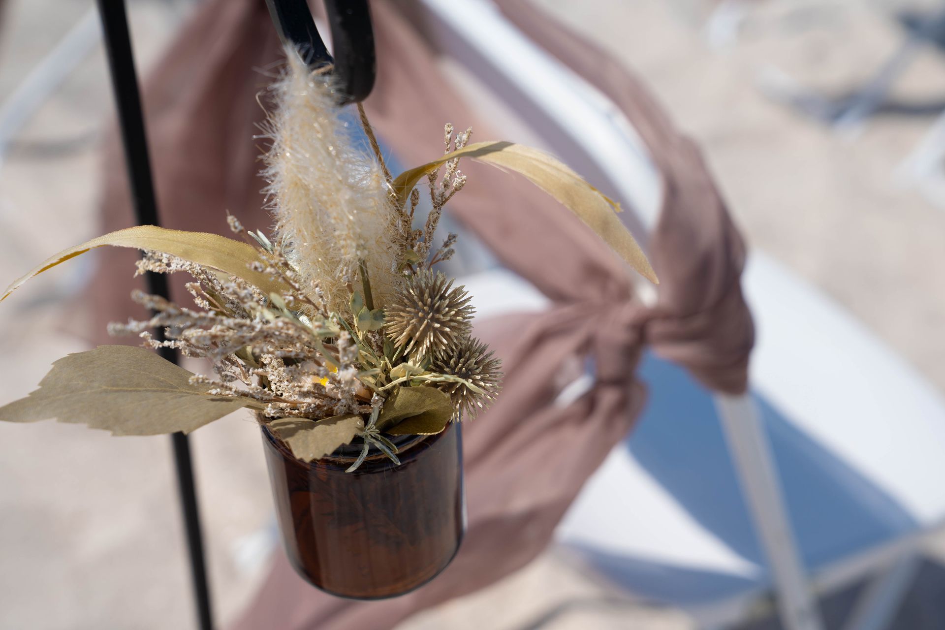 Floral arrangement in brown vase hanging on a black metal rod, with draped beige fabric and white chairs in the background.