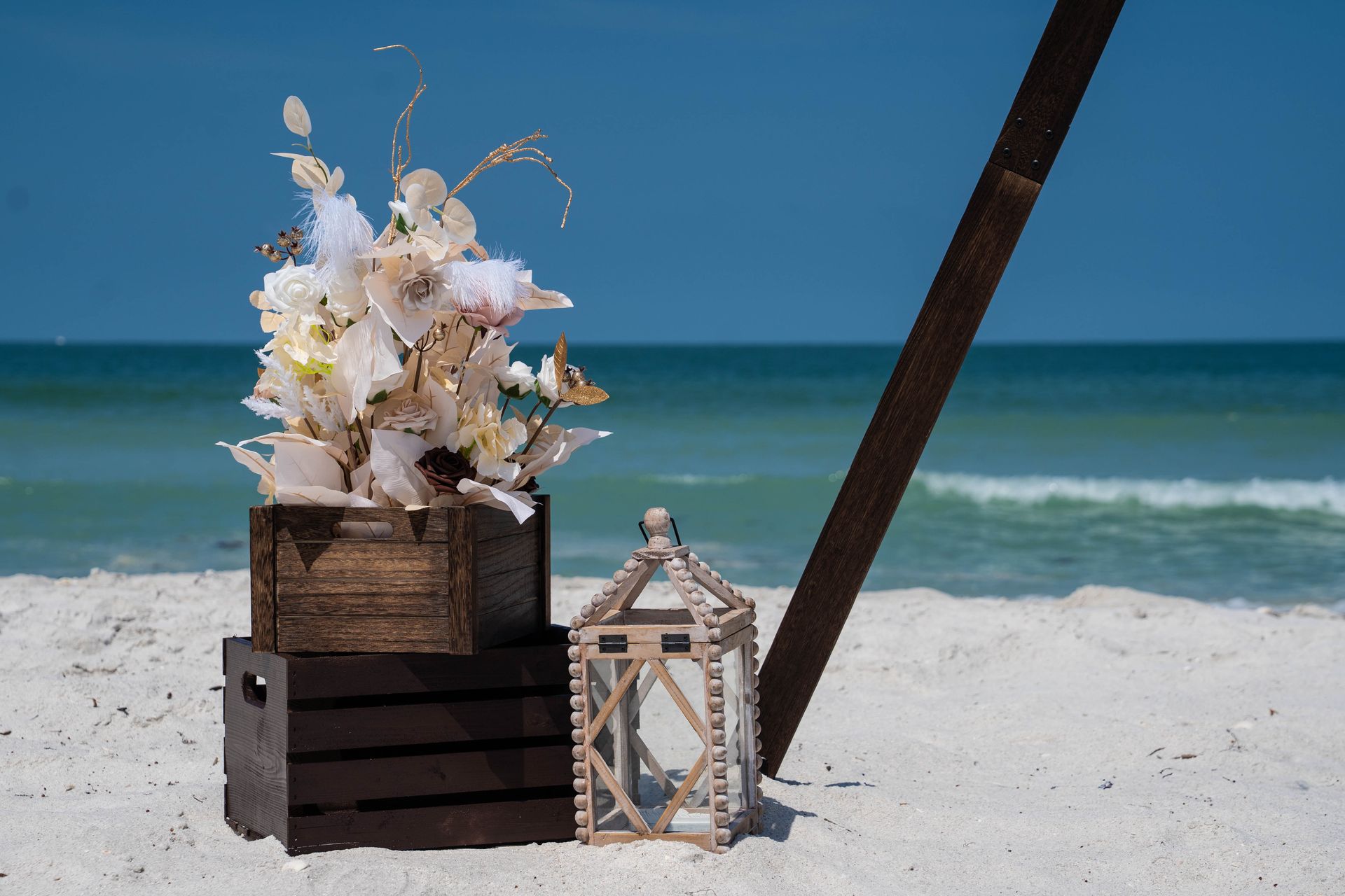 Beach wedding decor: floral arrangement atop crates, wooden lantern, blue ocean background.