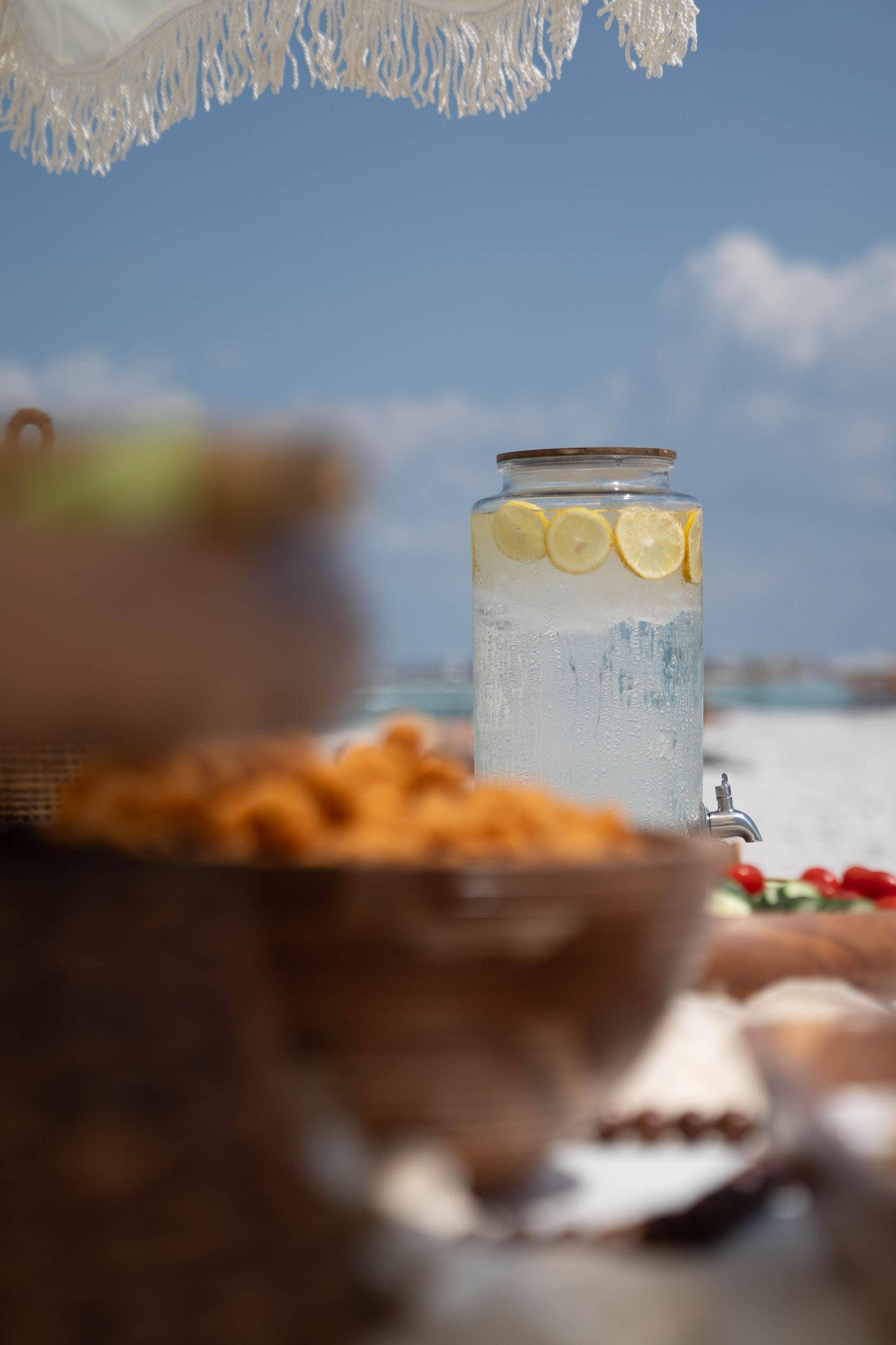 Lemon-infused water dispenser with sliced lemons, blurred food, and parasol against a blue sky.