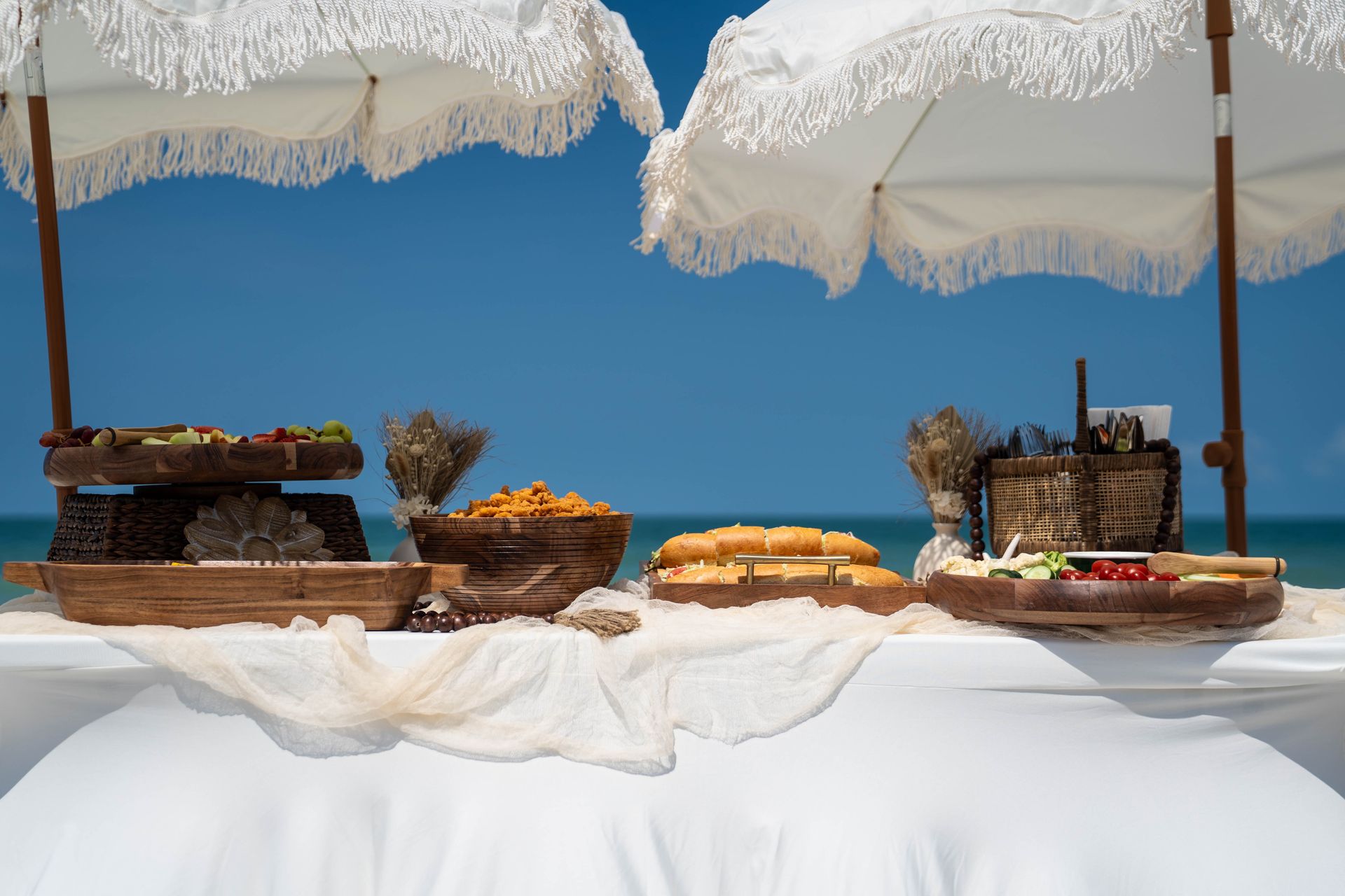 Beachside buffet table with food platters and parasols against a blue sky.