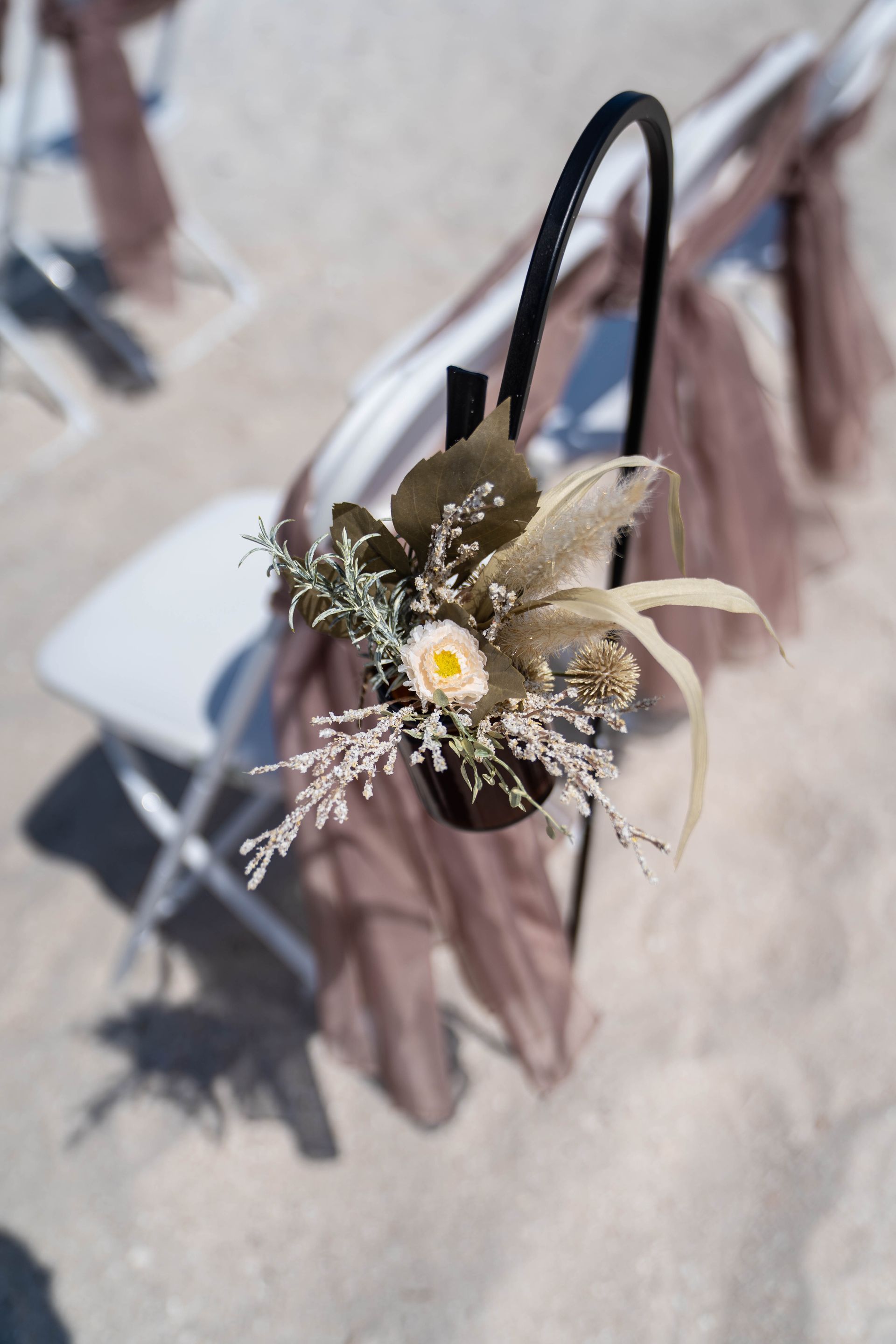 Close-up of a white folding chair with floral arrangement and draped fabric at a beach wedding.
