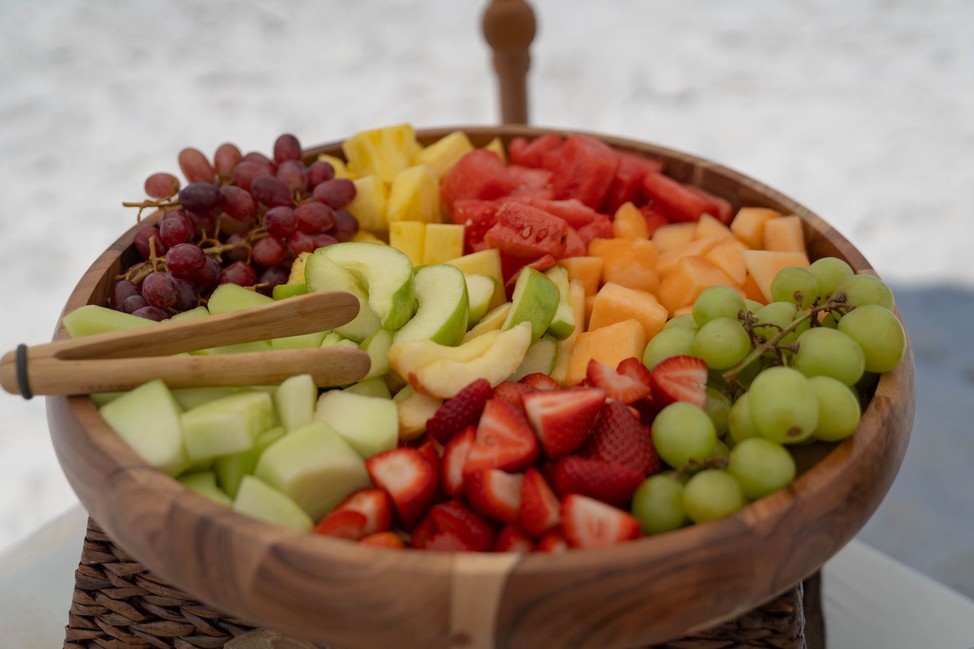 Wooden bowl filled with a rainbow of fresh fruit: grapes, melon, strawberries, apples, pineapple, on a white surface.