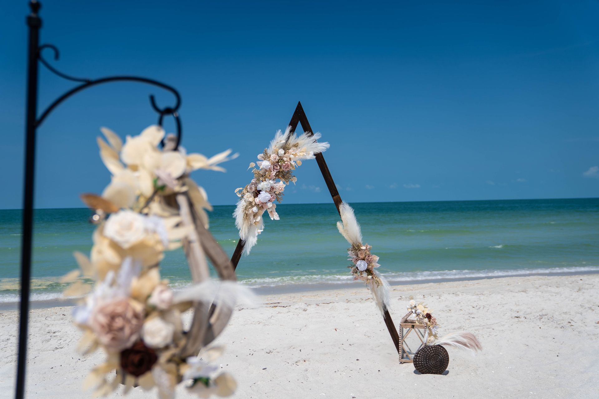 Beach wedding decor: floral arch on white sand, ocean backdrop, blue sky.