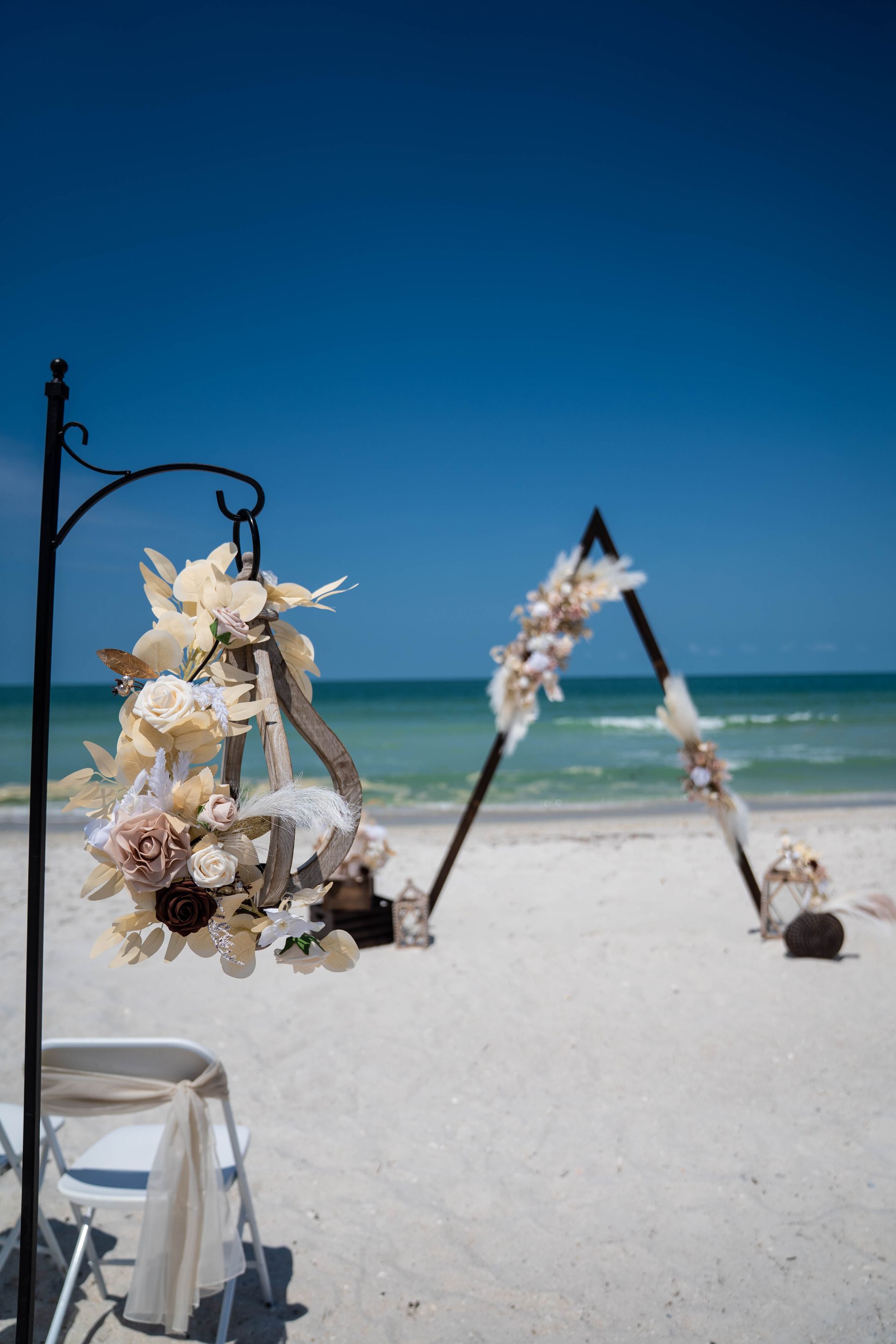 Beach wedding ceremony setup: triangle arch, floral decorations, white sand, and blue ocean.