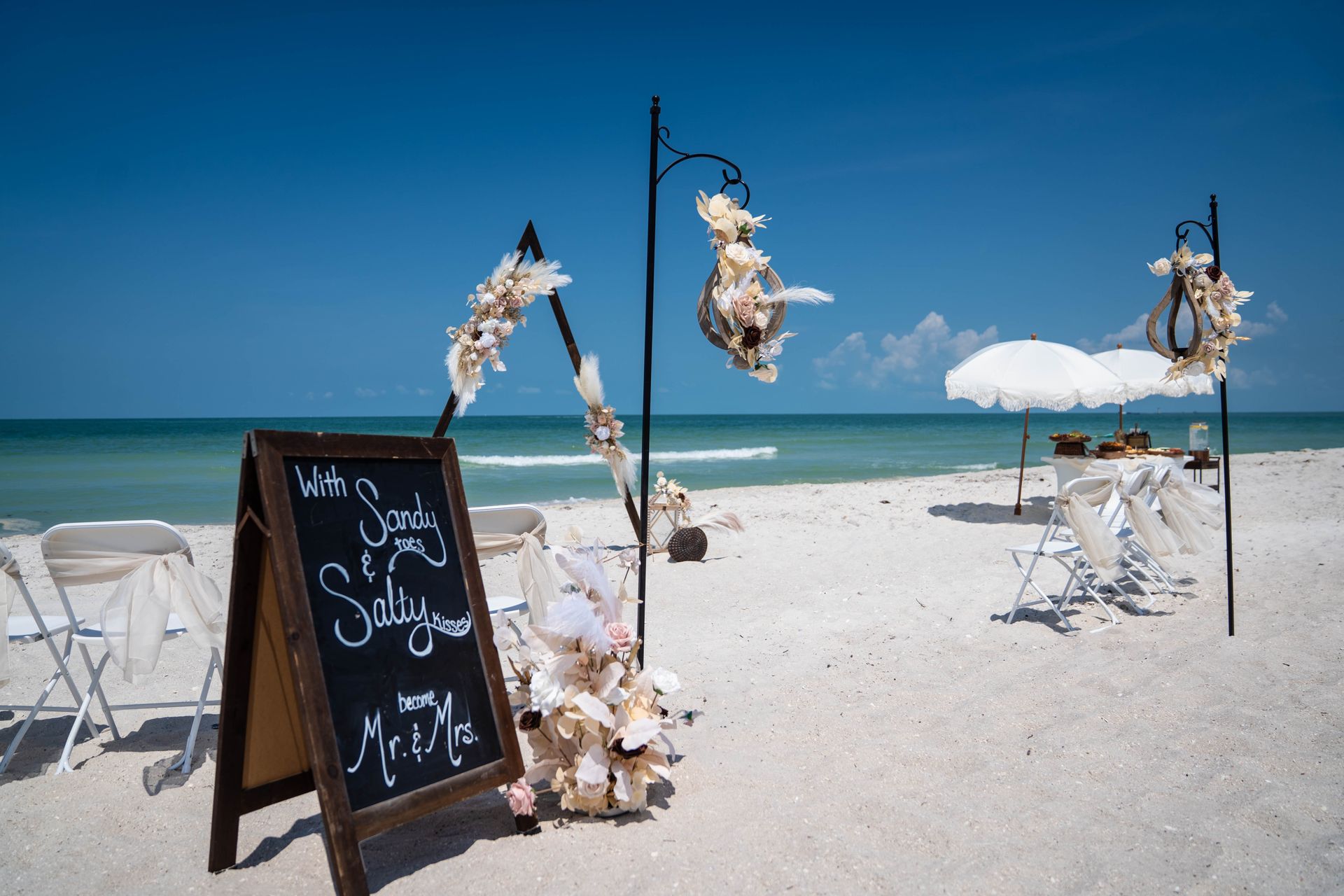 Beach wedding setup with a sign, chairs, and decorations under a clear blue sky.
