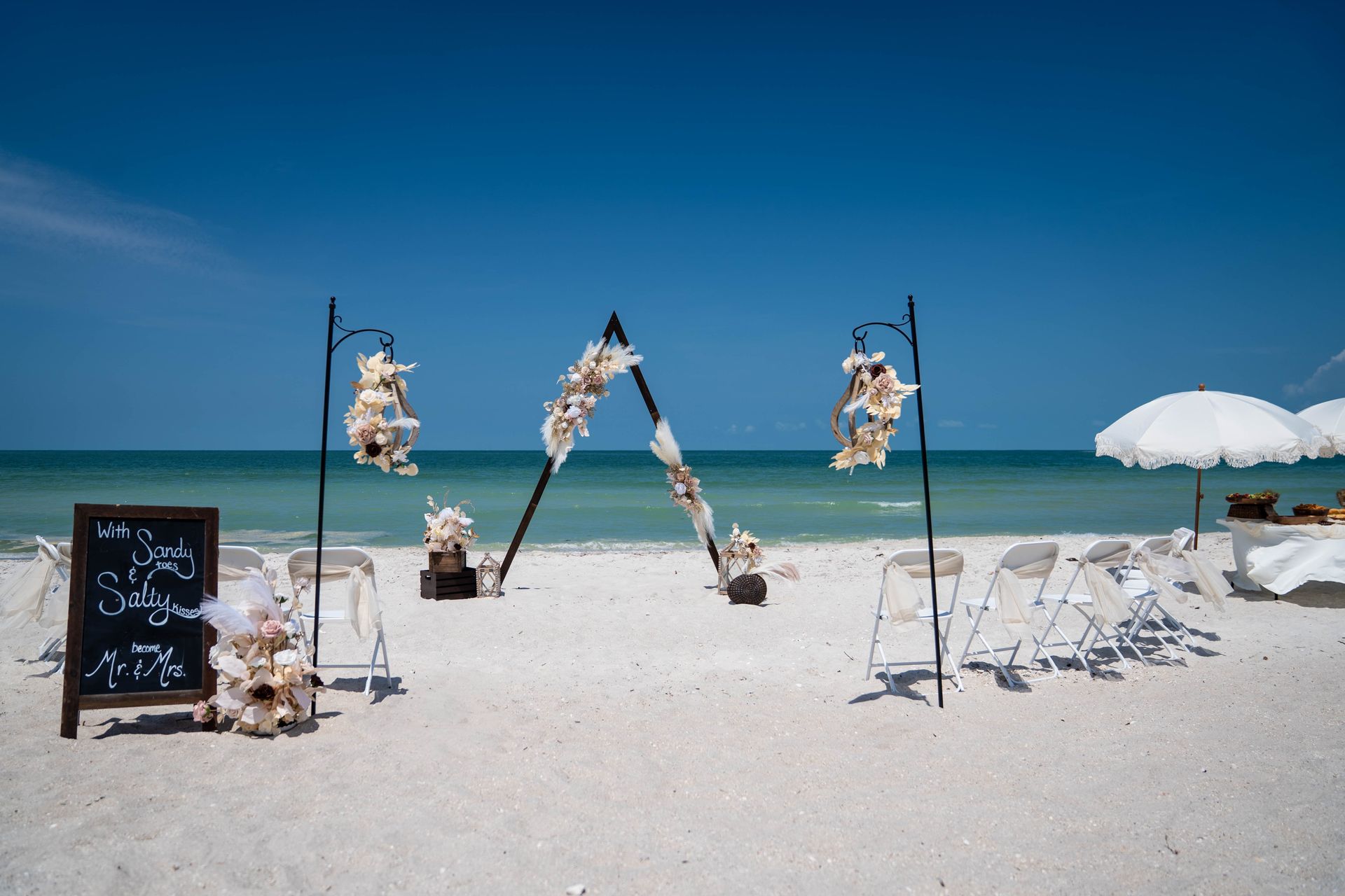 Beach wedding ceremony setup: arch with shell decorations, chairs, sign on sand, bright blue sky.